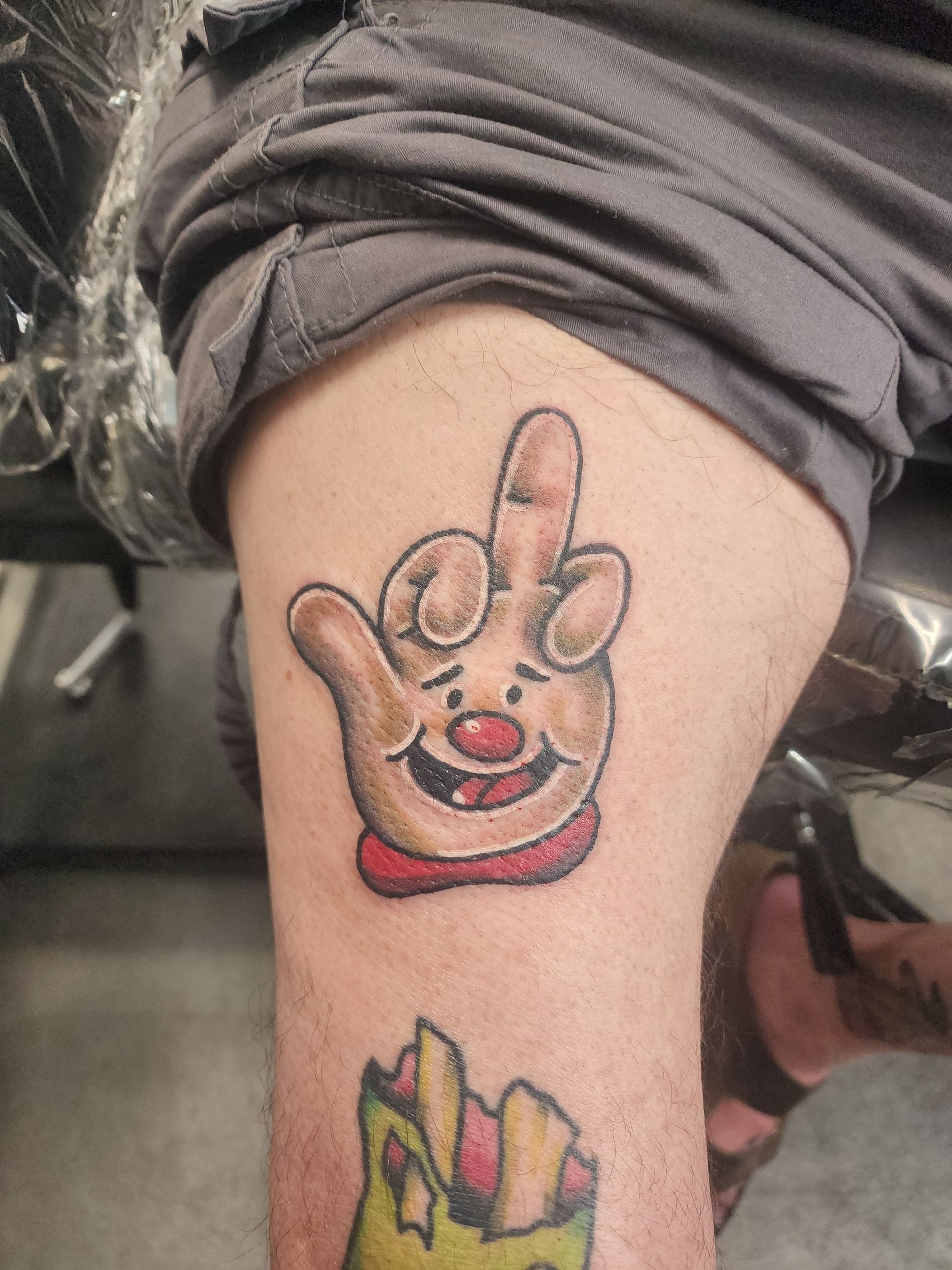 Tattoo of a cartoon hand with a raised middle finger, red nose, and smile, on a leg.