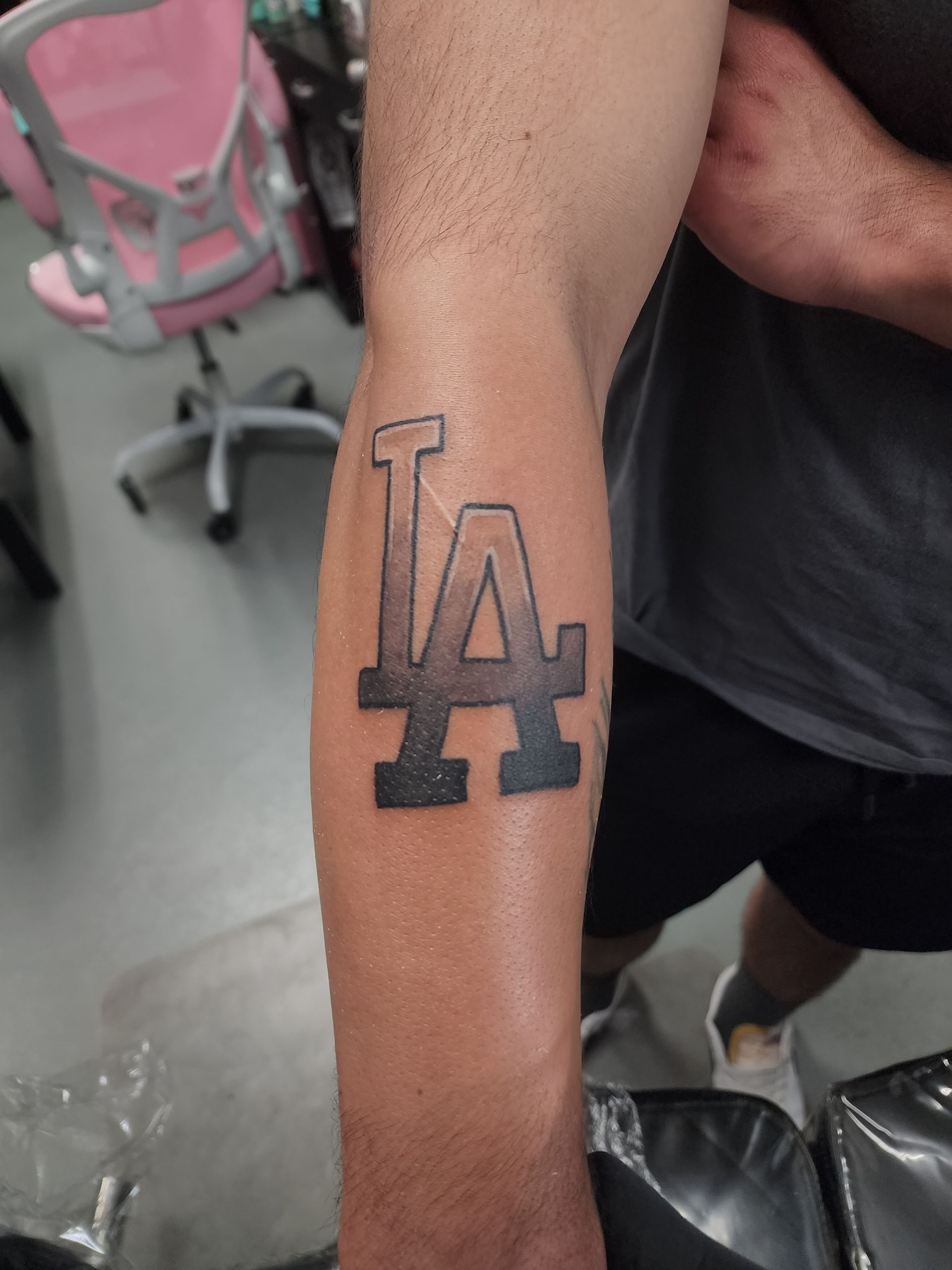 Tattoo of LA Dodgers logo on a person's forearm. Black and gray, indoor setting.