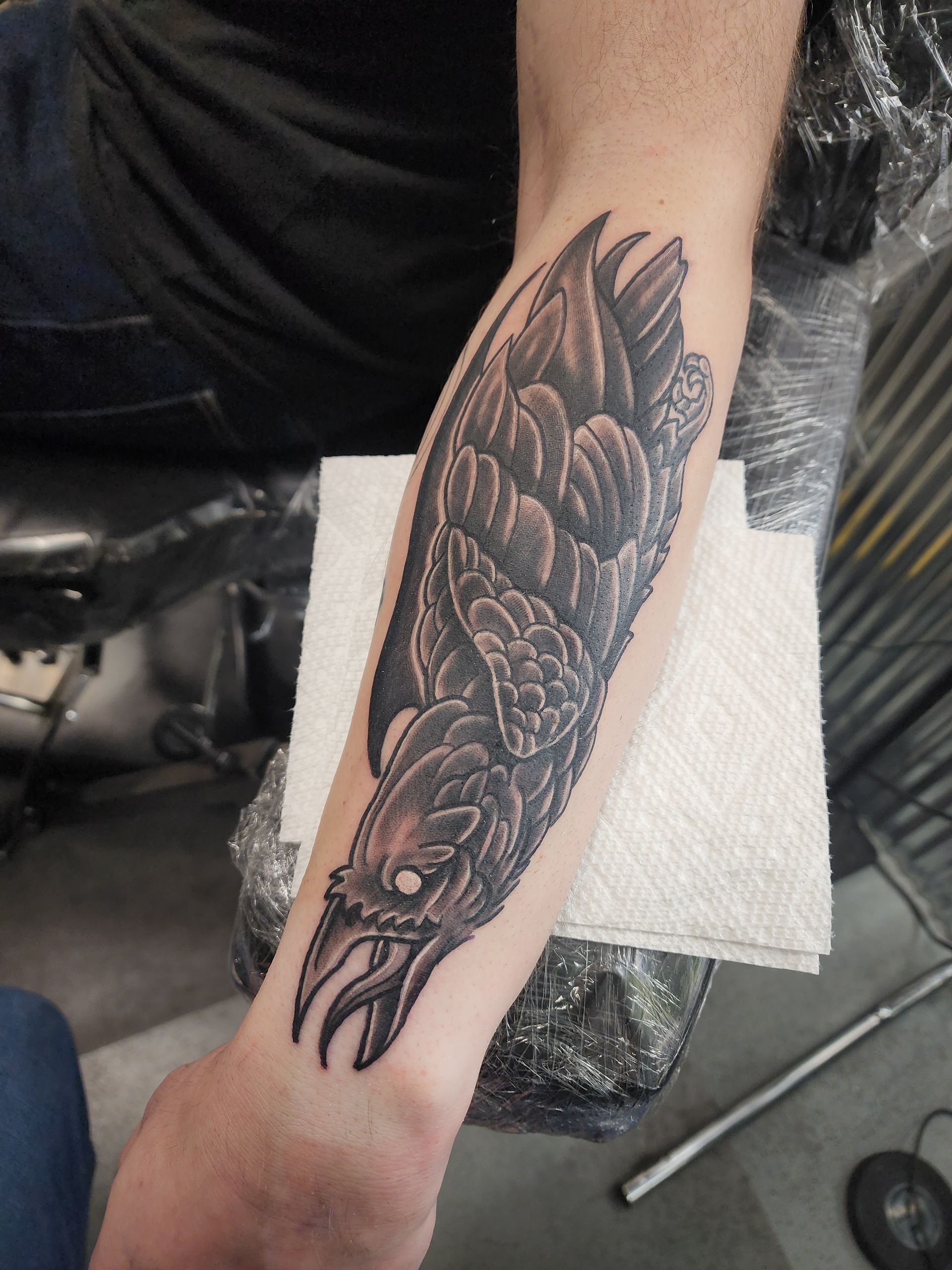 Arm with tattoo of a dark, stylized bird. It's partially wrapped in plastic, on a tissue.