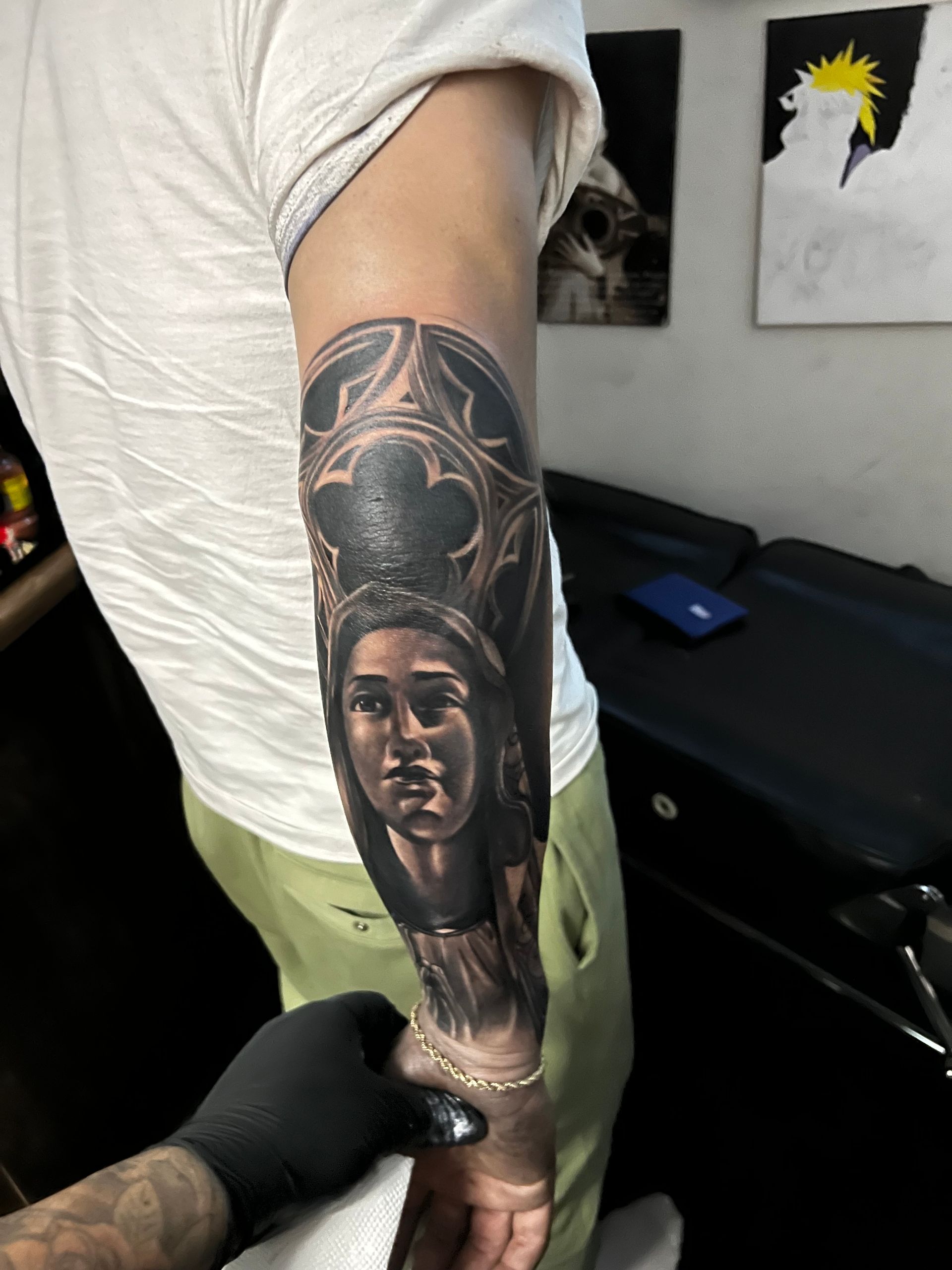 Person's arm with a detailed gothic-style portrait tattoo. Gloved hand holds the arm during the process.