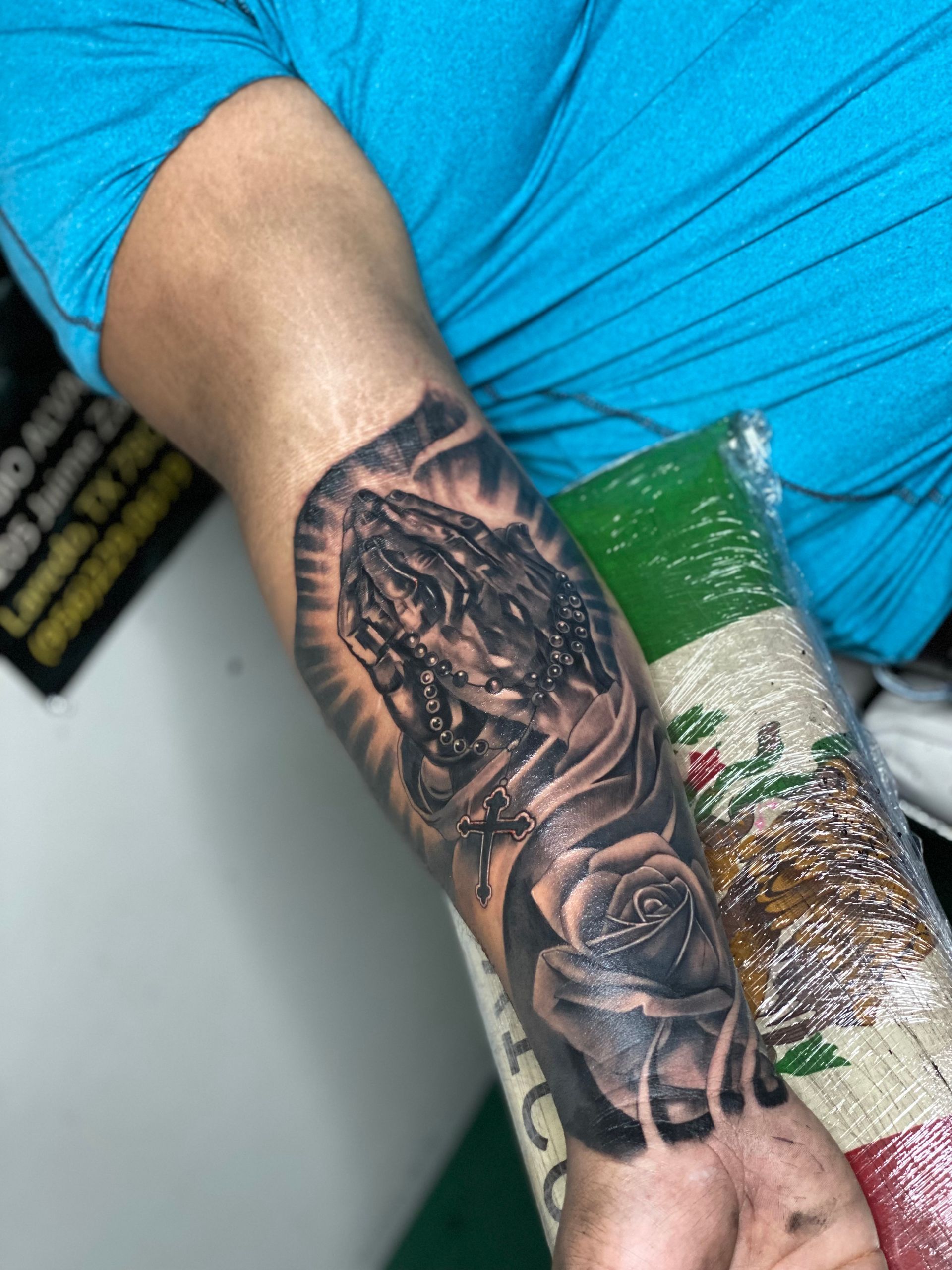 Tattoo of praying hands and a rose on a person's forearm, in grayscale.