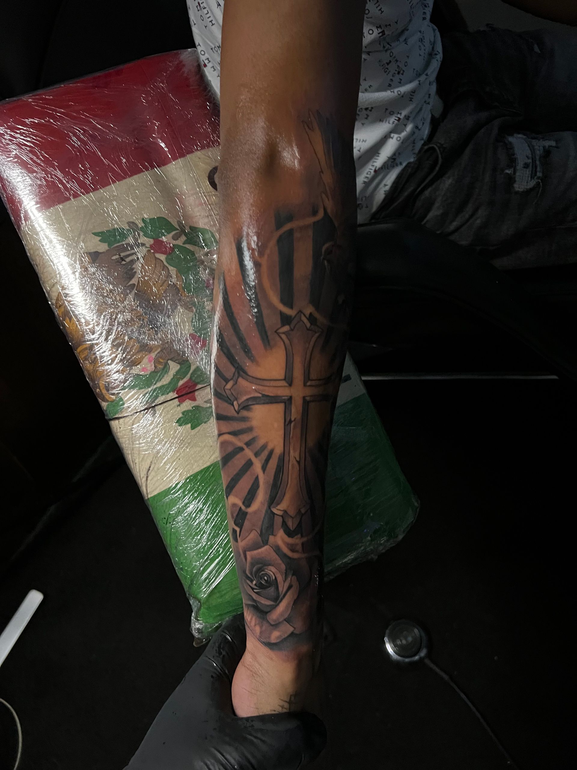 Arm tattoo: Cross with rays, roses, and initials. Arm is held with a Mexican flag in the background.
