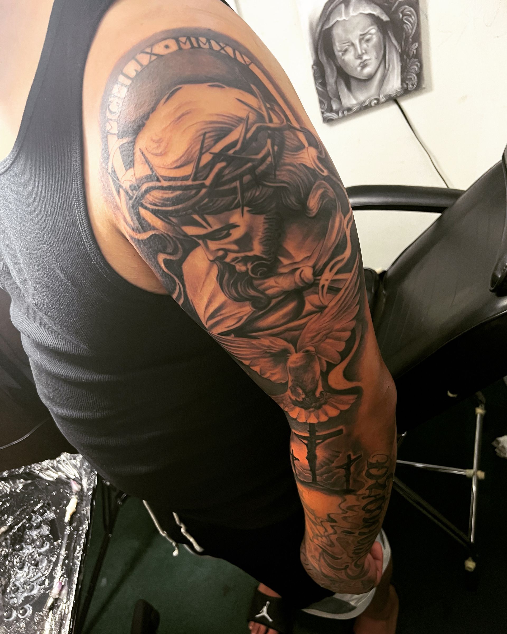 Tattoo on arm depicting Jesus with crown of thorns, dove, and cross in black and gray.