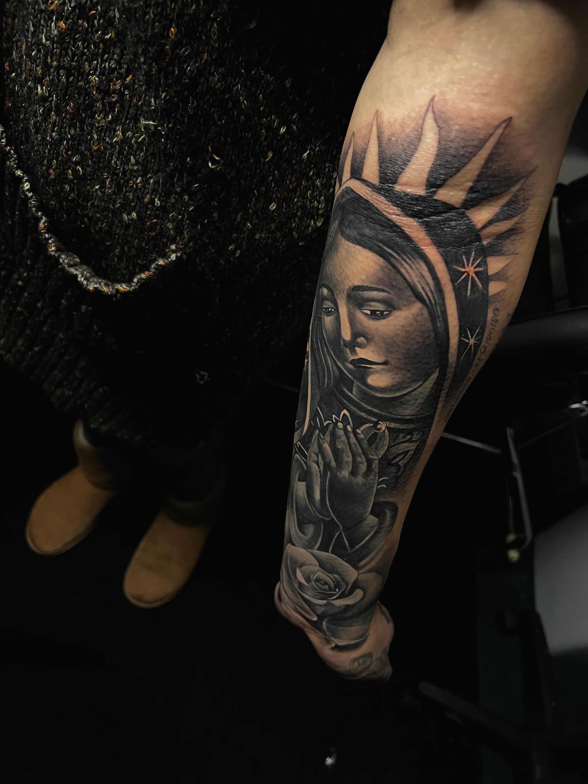 Black and white tattoo of Virgin Mary on arm, praying, with halo and rose.