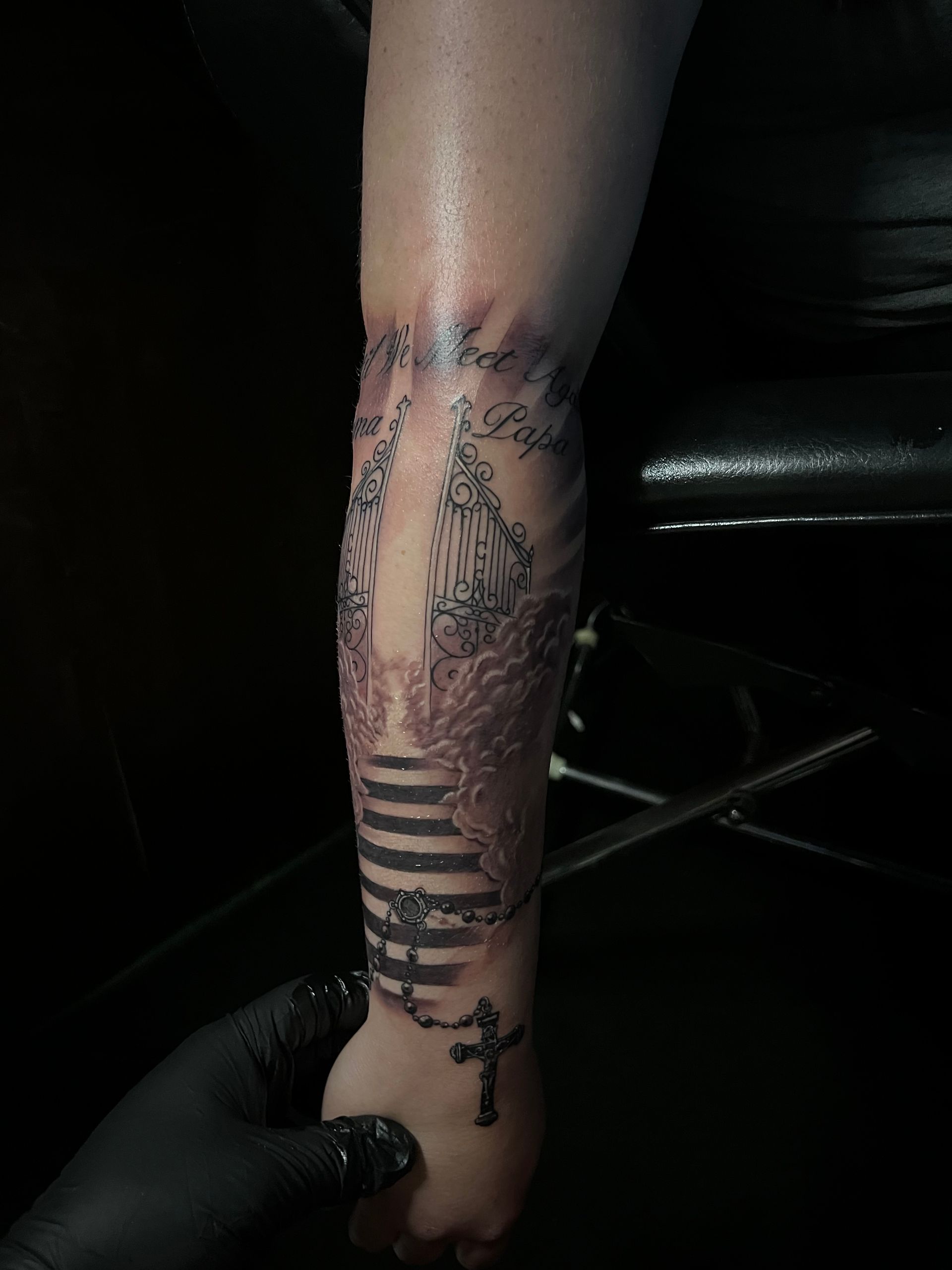 Tattoo on forearm; stairs design with rosary and cross on wrist. Dark setting, black gloved hand holding arm.