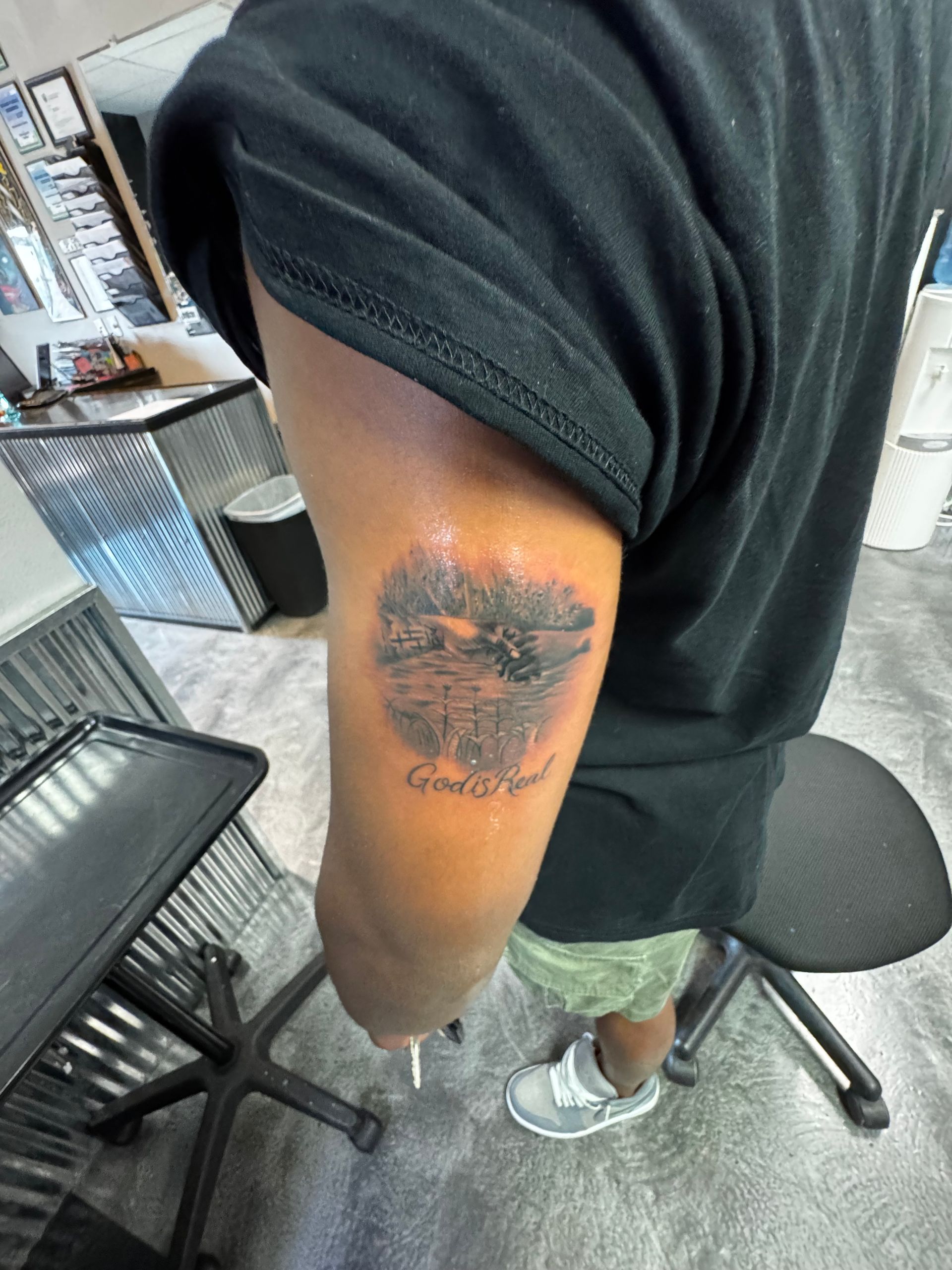 Person's arm with a tattoo of a lake scene, dark clothing, and a tattoo shop setting.