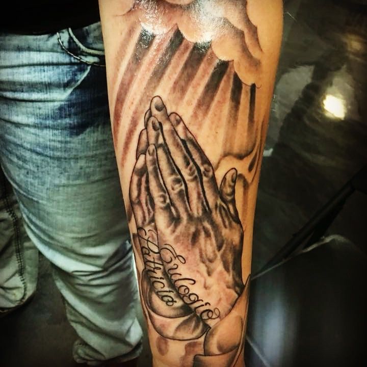 Tattoo of praying hands with light rays beaming down from the clouds, and writing: