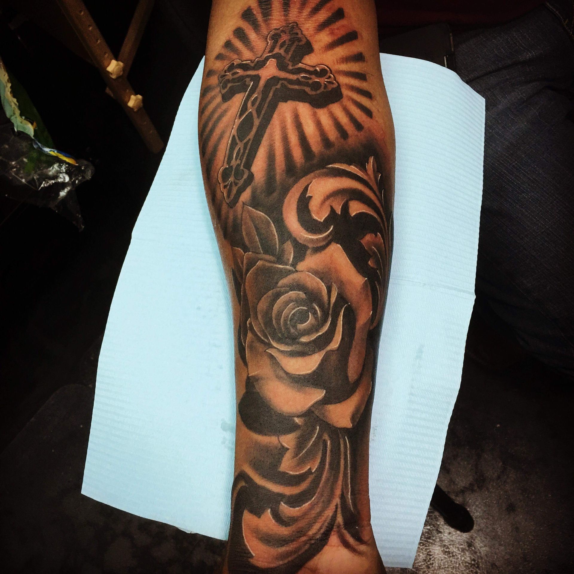 Arm tattoo of a cross with rays, a rose, and swirling designs, all in grayscale.