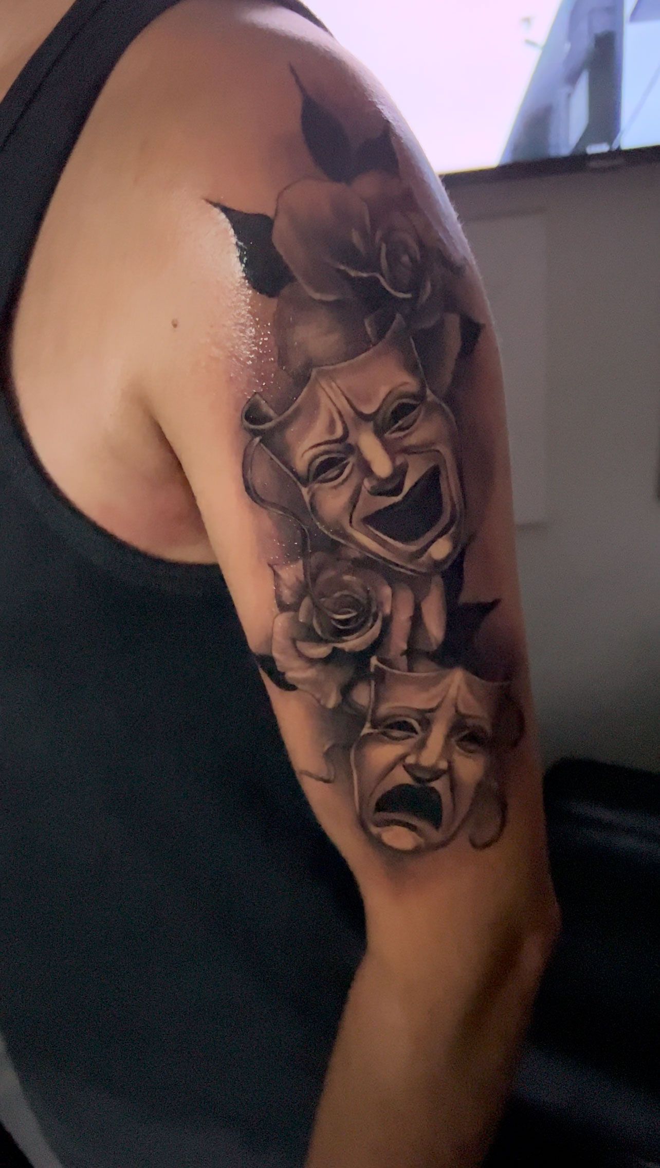 Tattoo of comedy and tragedy masks with roses on a person's upper arm. Gray and black.