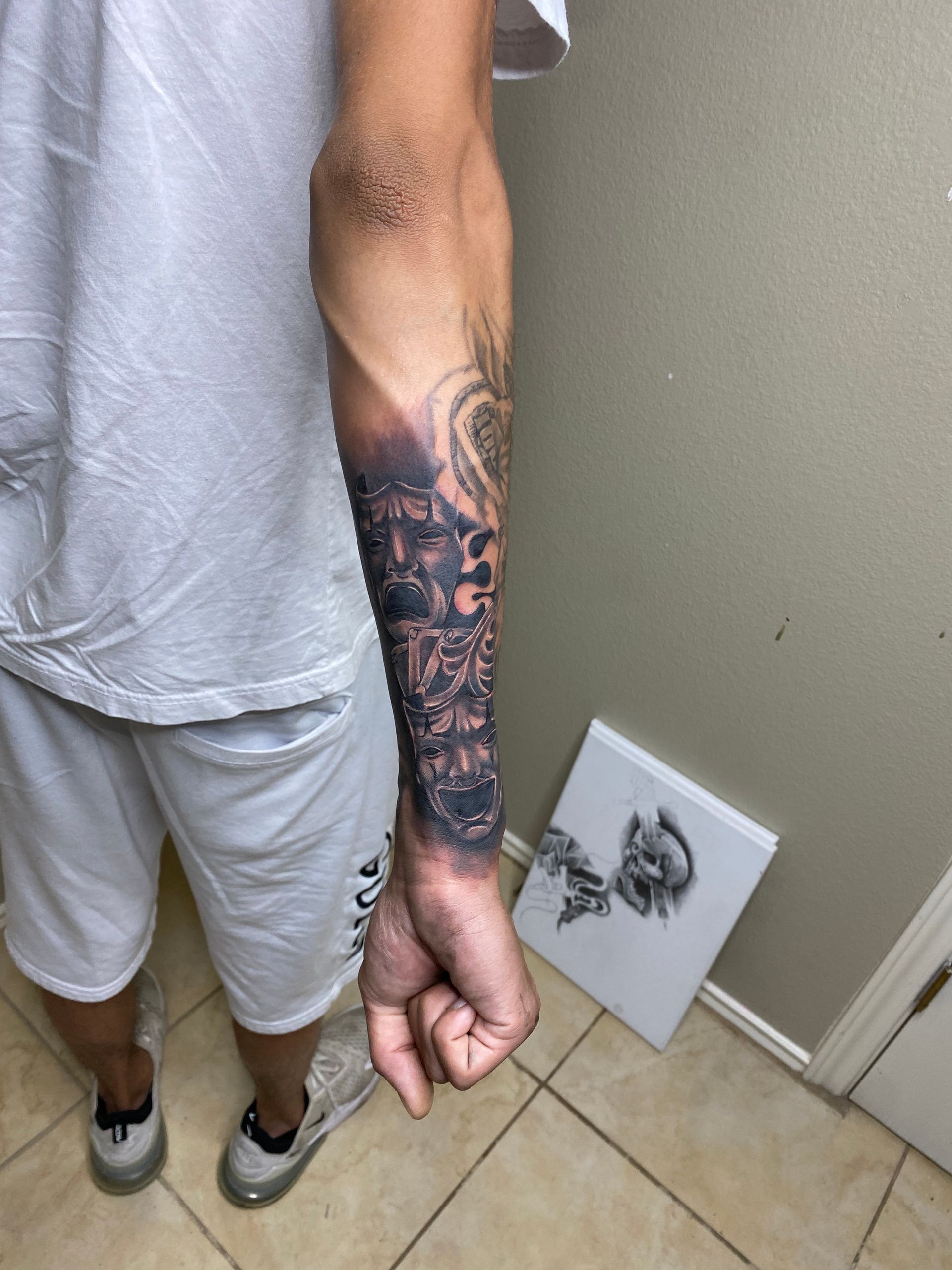 Person with a detailed forearm tattoo, wearing white clothing and sneakers, standing indoors.