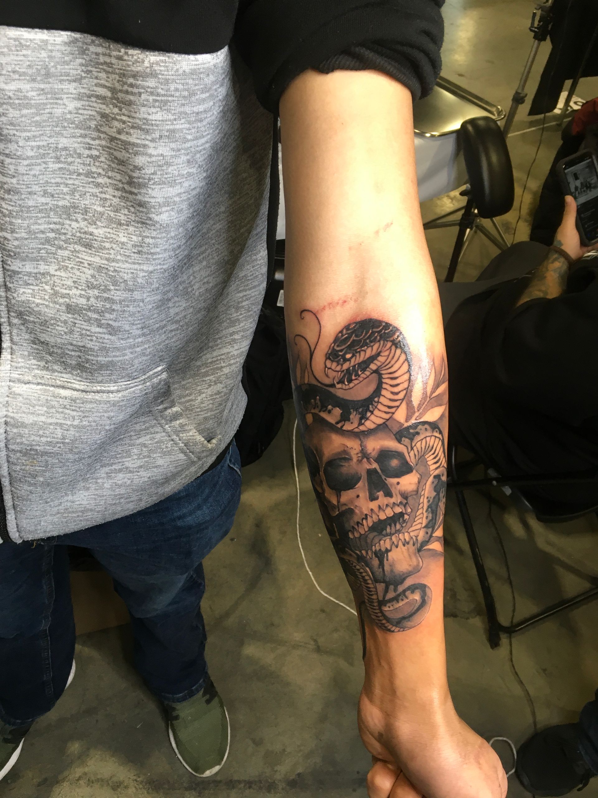 Tattoo of a skull and snake on a person's forearm. Black and gray. Person wearing gray shirt and jeans.