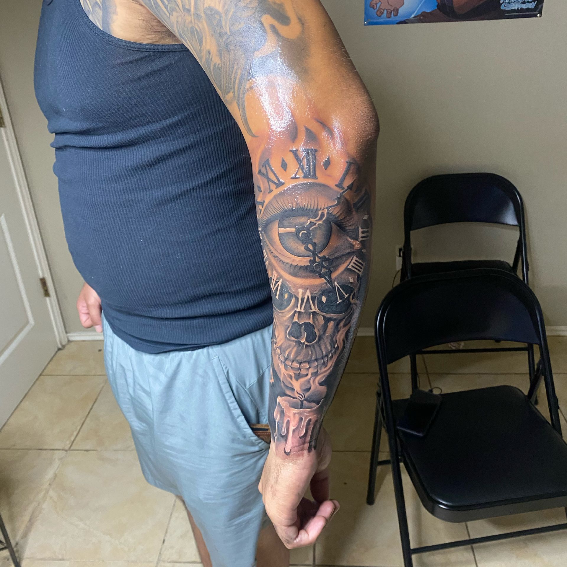 Man with forearm tattoo of eye, skull, and clock, wearing a black tank top and gray shorts.