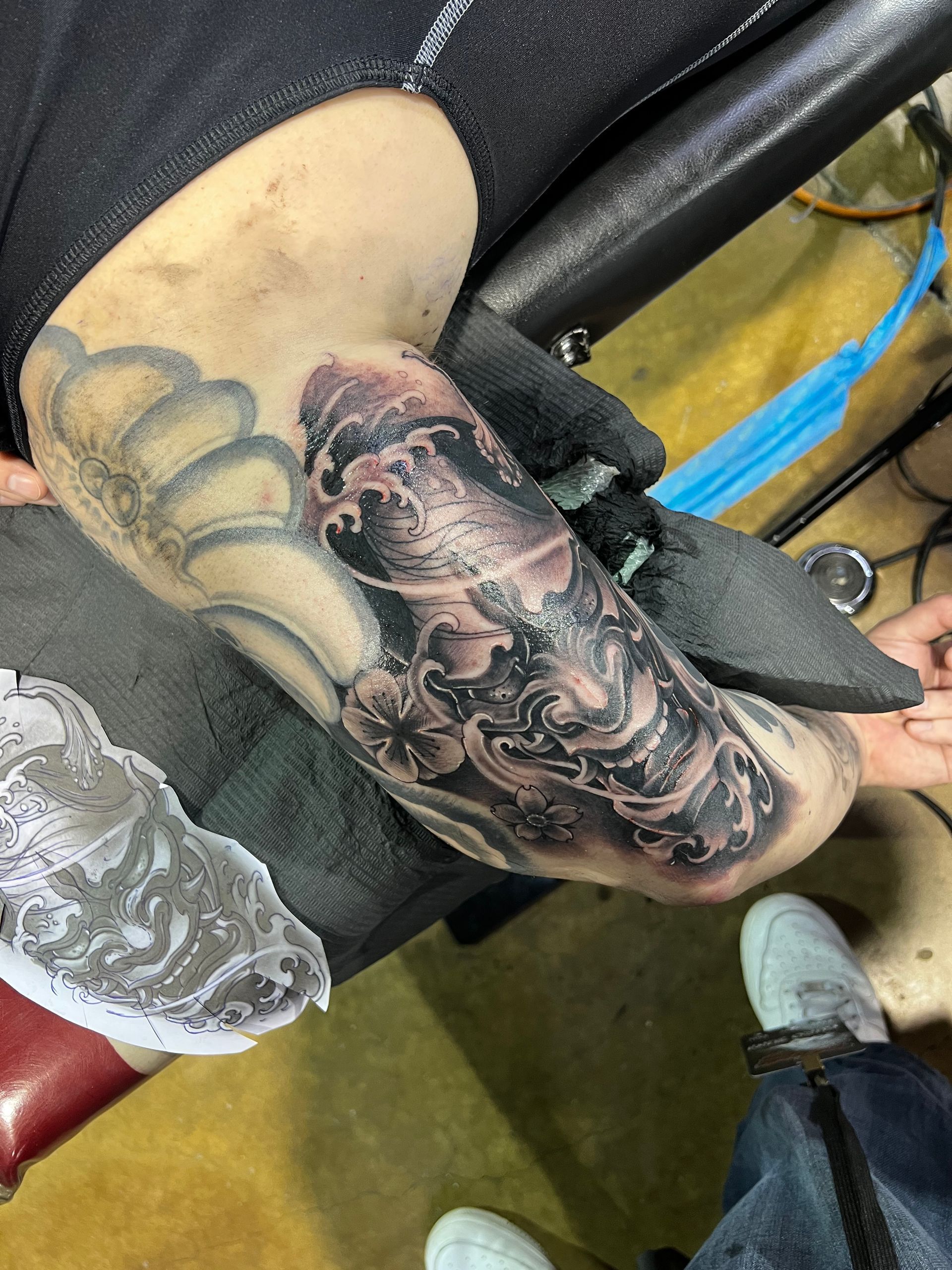 Tattoo artist working on a detailed, black and gray arm tattoo. Person's arm bent, black gloves visible.