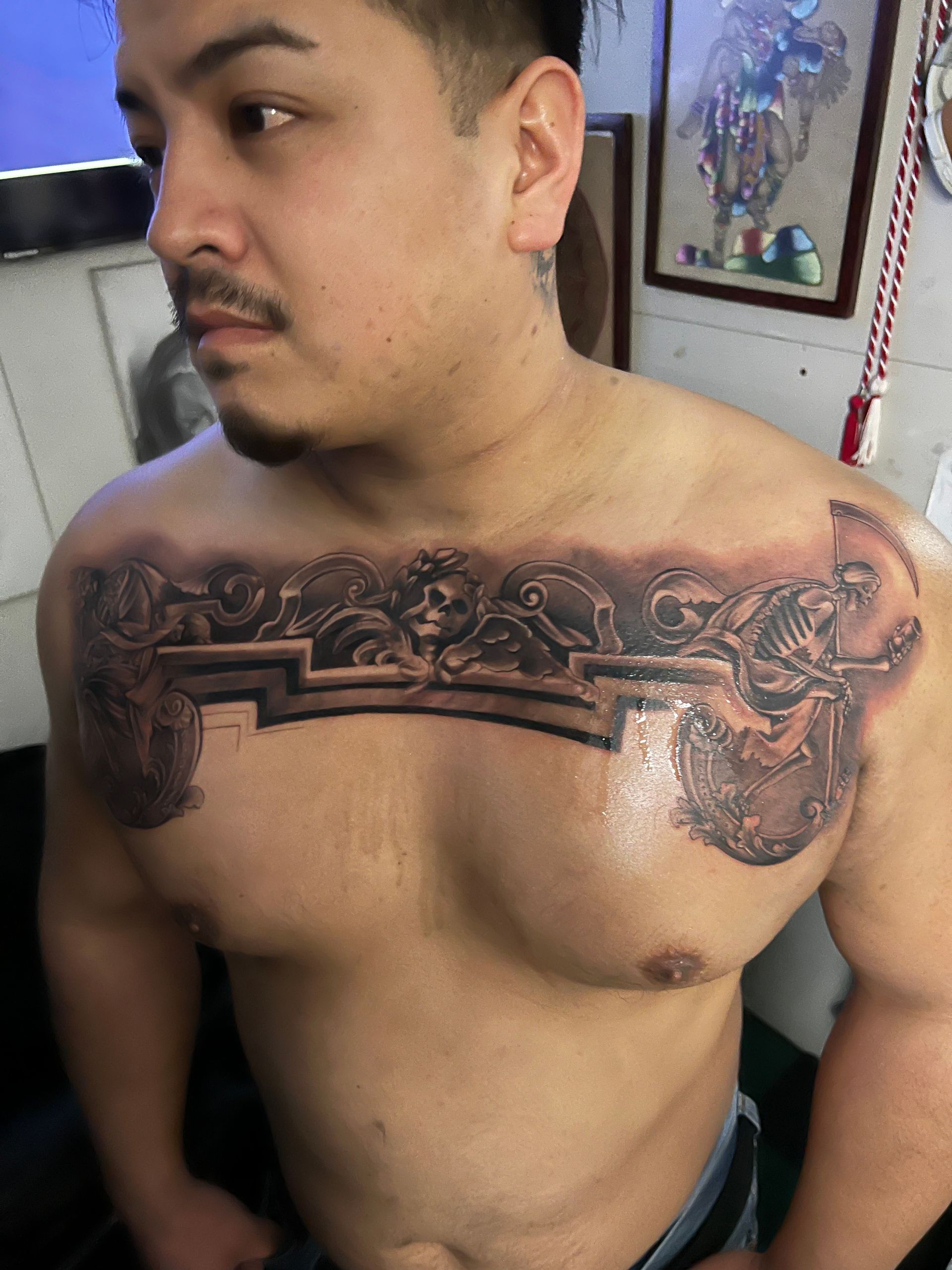 Man with chest tattoo; dark gray intricate design with skulls, arch, and angel figure. Indoor setting.