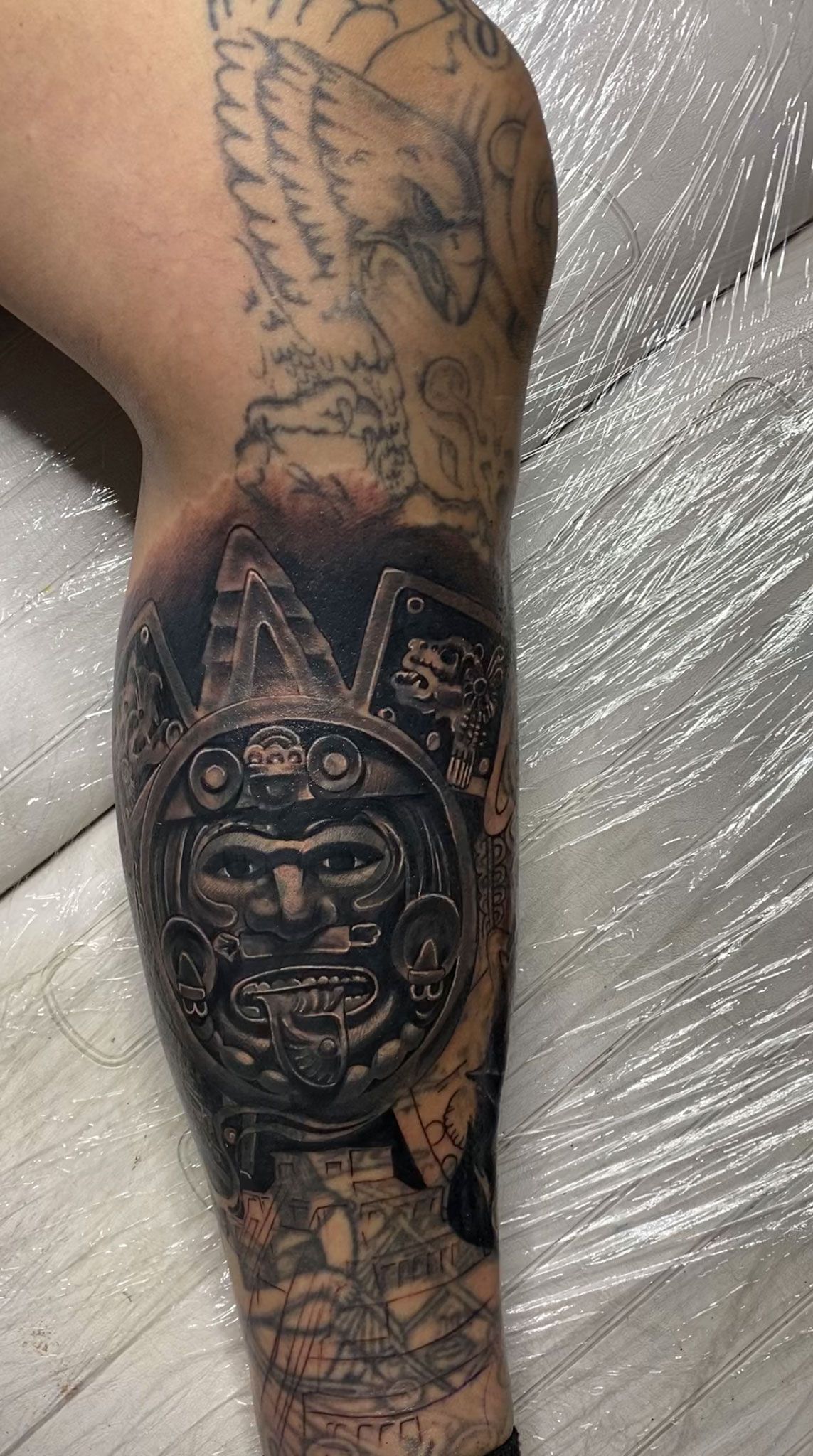 Aztec sun stone tattoo on a leg; detailed, dark shading, surrounded by other faded tattoos.