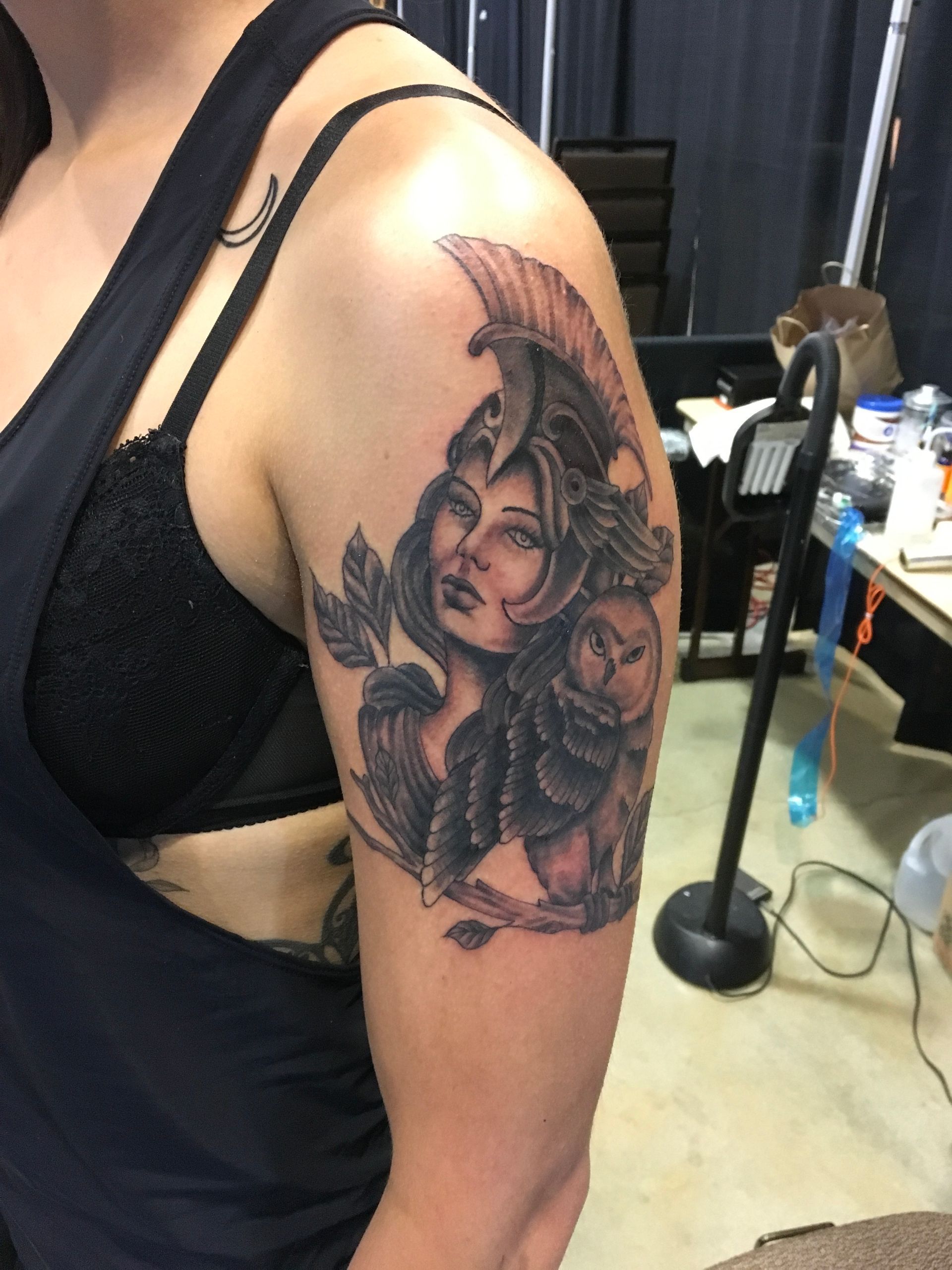 Woman's arm with tattoo of Athena in helmet, owl, and leaves; black and grey, indoors.