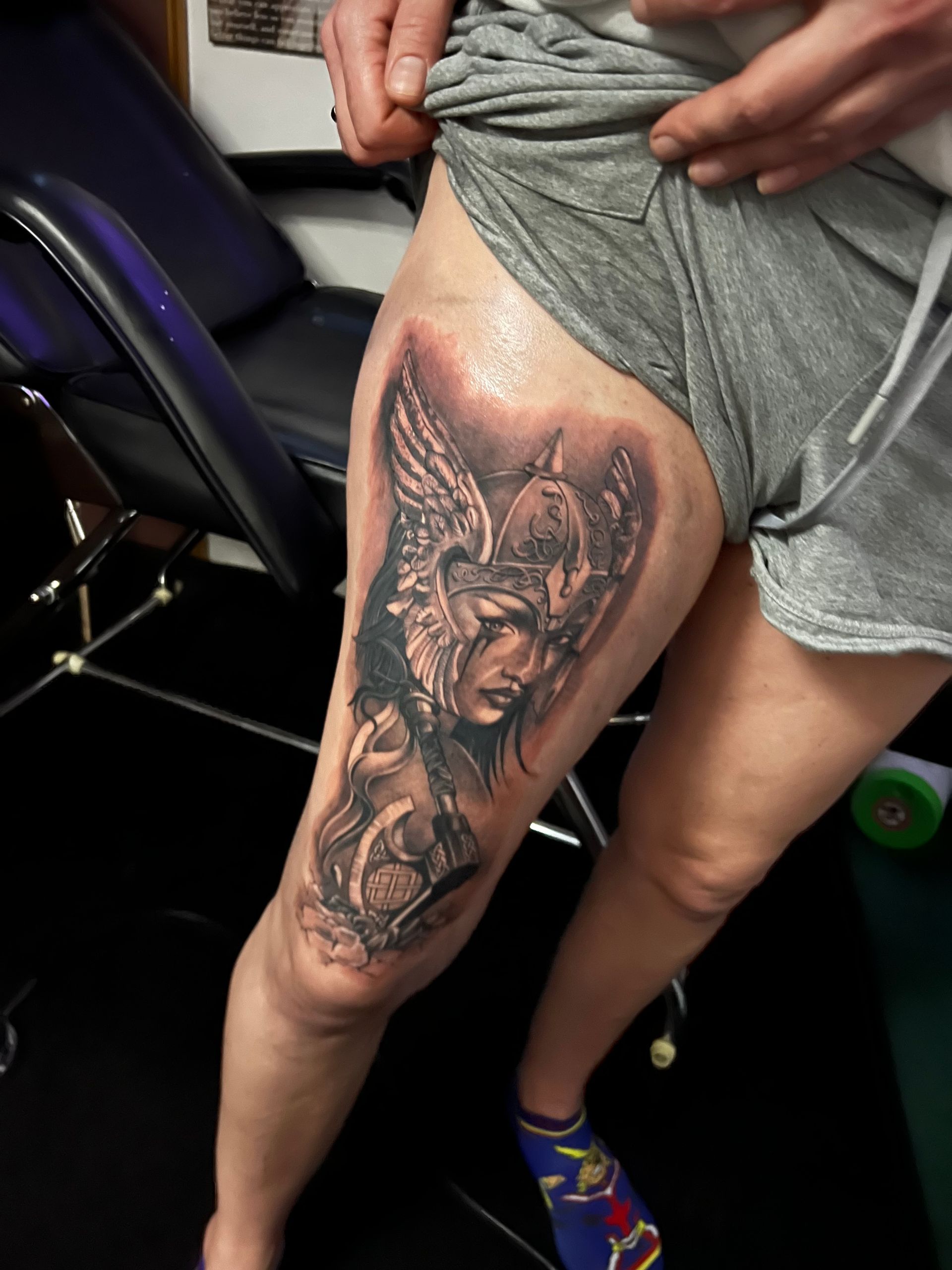 Tattoo of a woman in armor with wings on a person's thigh. Black, grey, and white design in a tattoo parlor.