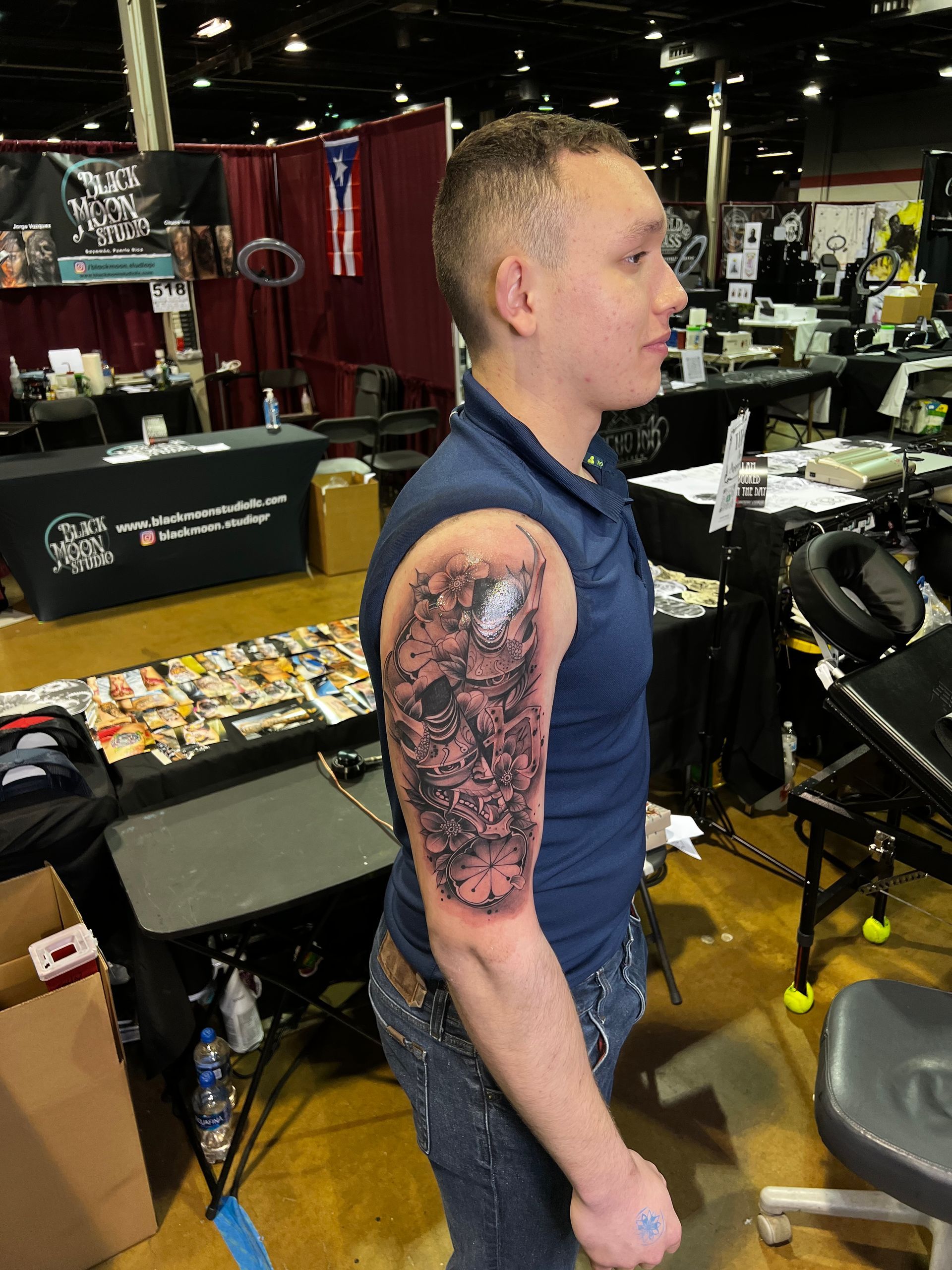 Man with a large, dark arm tattoo stands at an event booth. He wears a blue shirt and jeans, facing right.