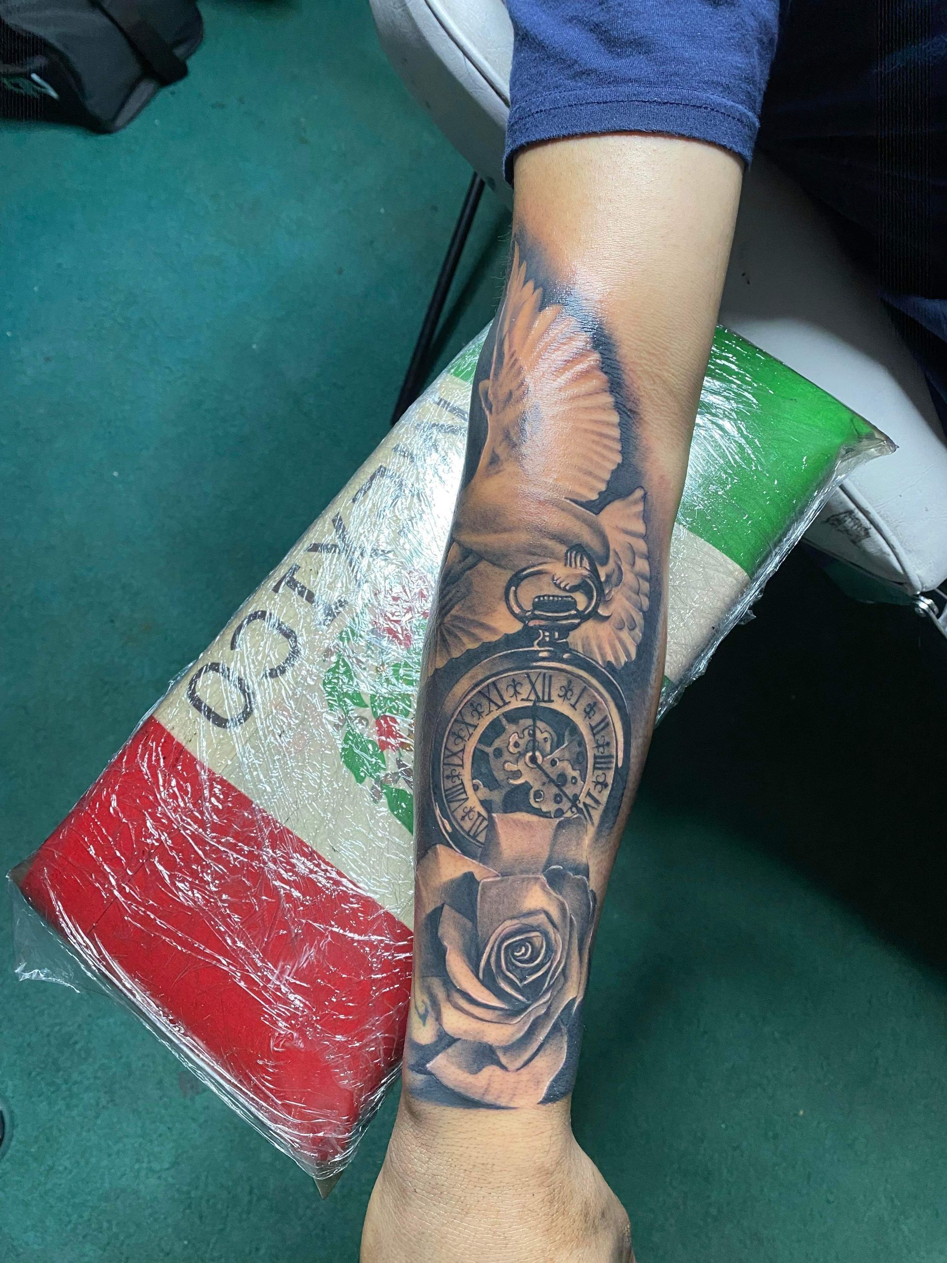Tattoo of a pocket watch, dove, and rose on a person's arm. The arm is resting on a Mexican flag.