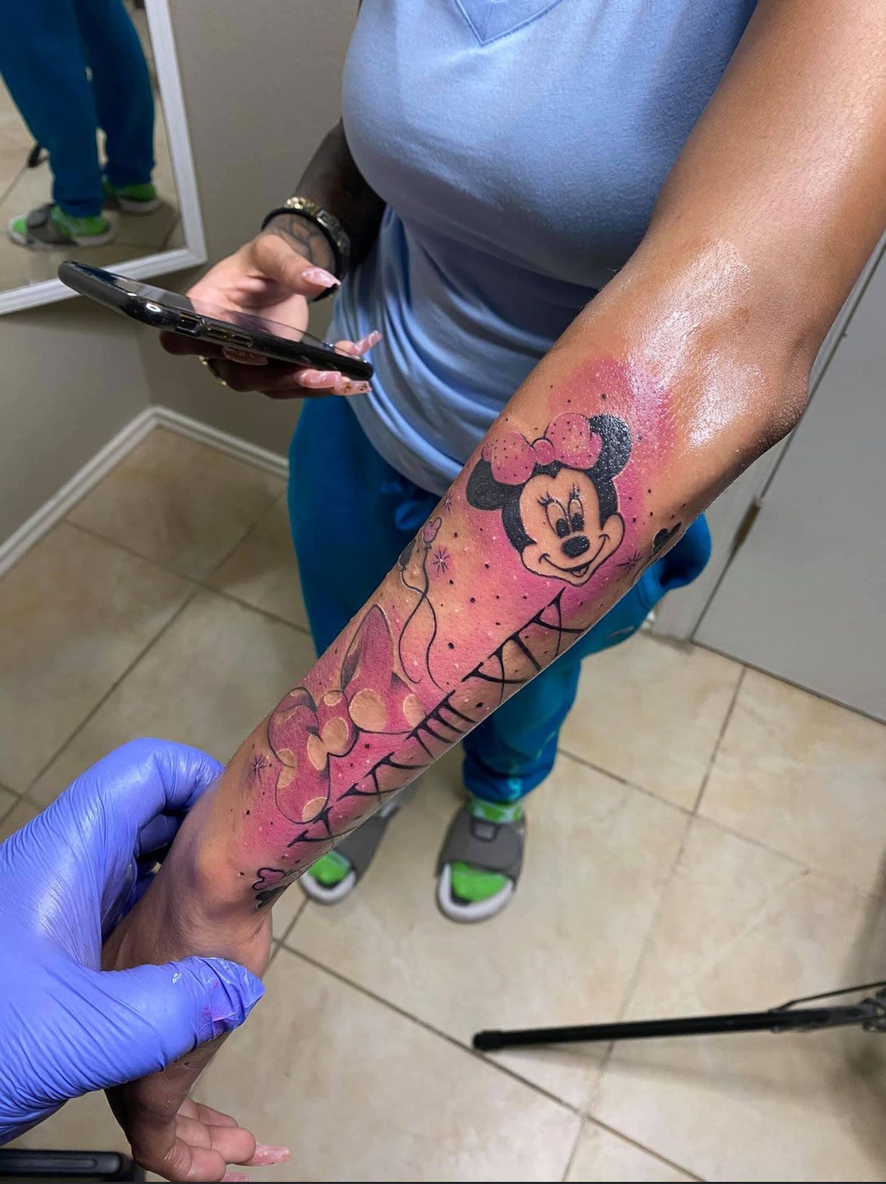 Person's arm with fresh Minnie Mouse tattoo, pink, black, and numbers. Others in the background.