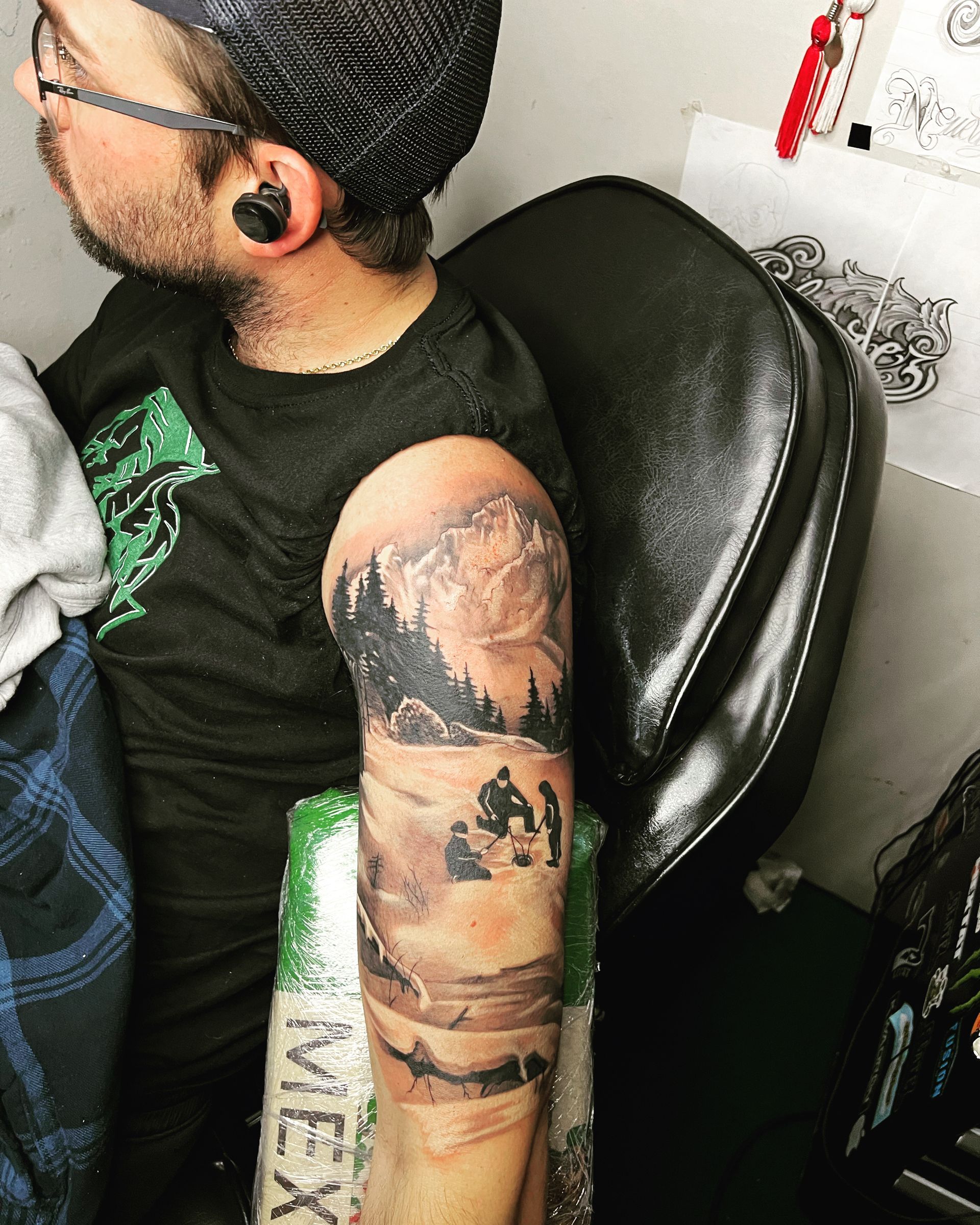 Person with arm tattoo depicting a landscape scene with mountains, trees, and figures.