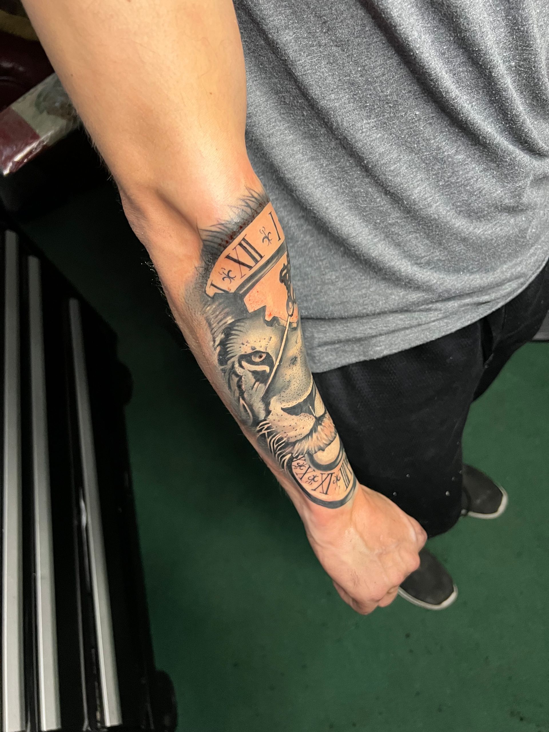 Tattoo of a clock, skull, and gears on a person's forearm. Black and gray with a light orange backdrop.