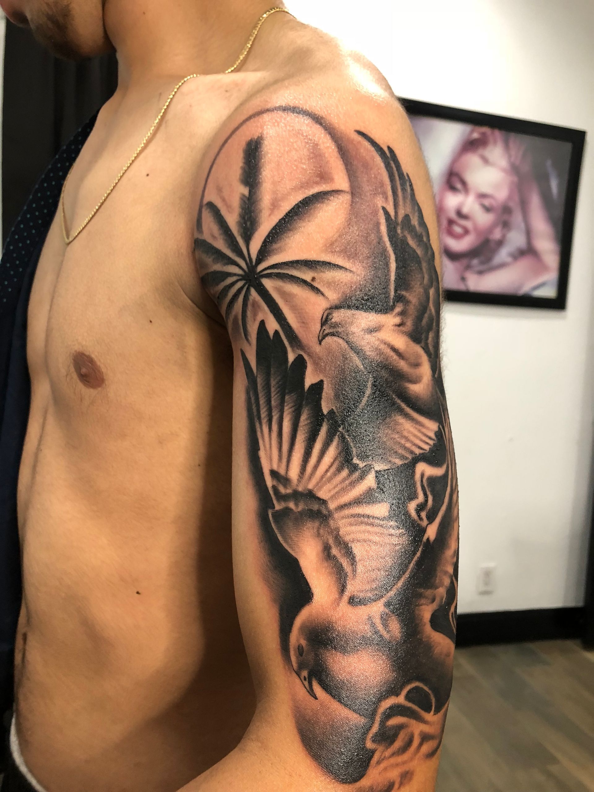 Tattoo of two doves flying on a man's upper arm. One dove is in front of a palm tree. In the background, there's a photo of Marilyn Monroe.