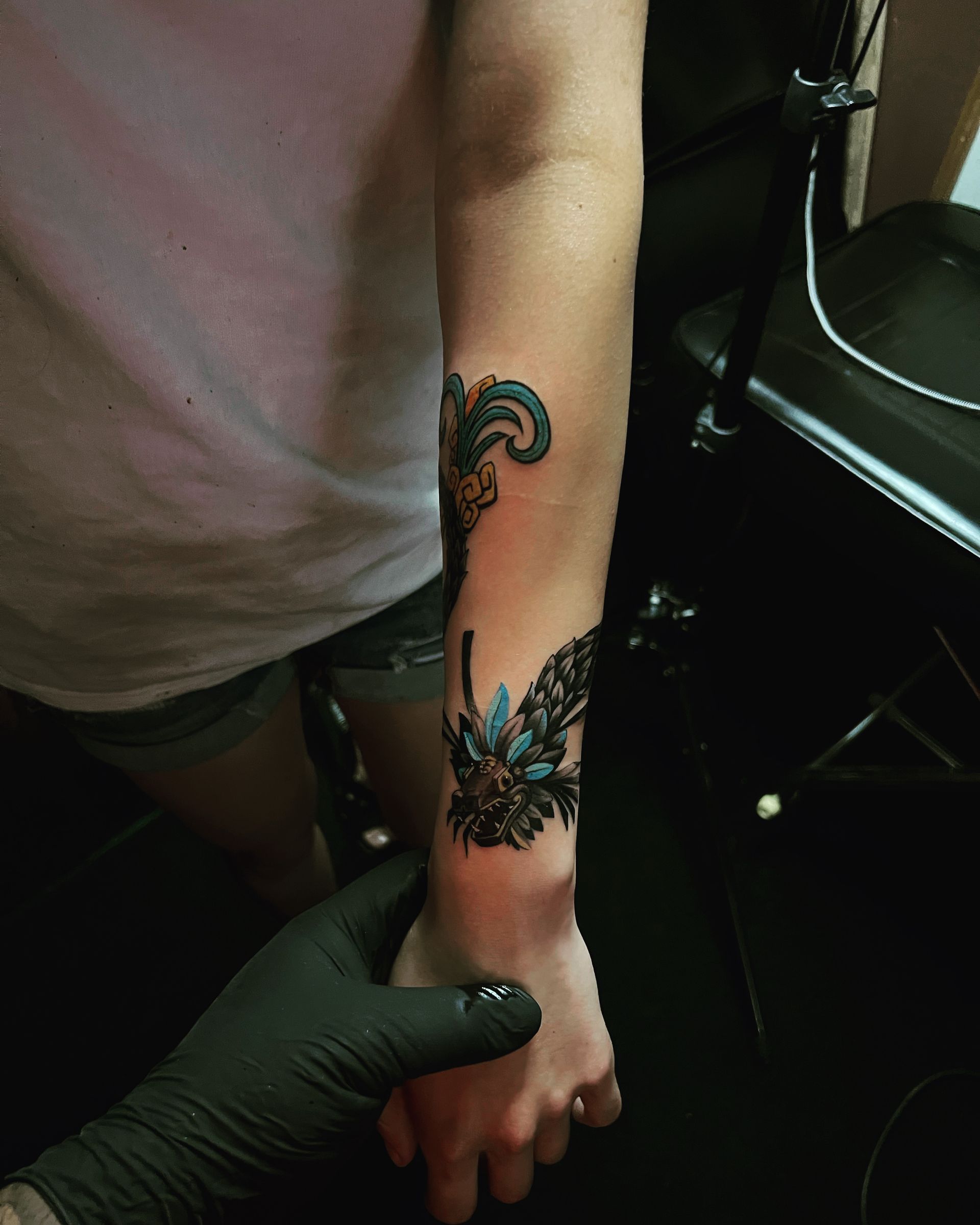Arm with teal and black tattoo, held by a gloved hand in a dark setting.