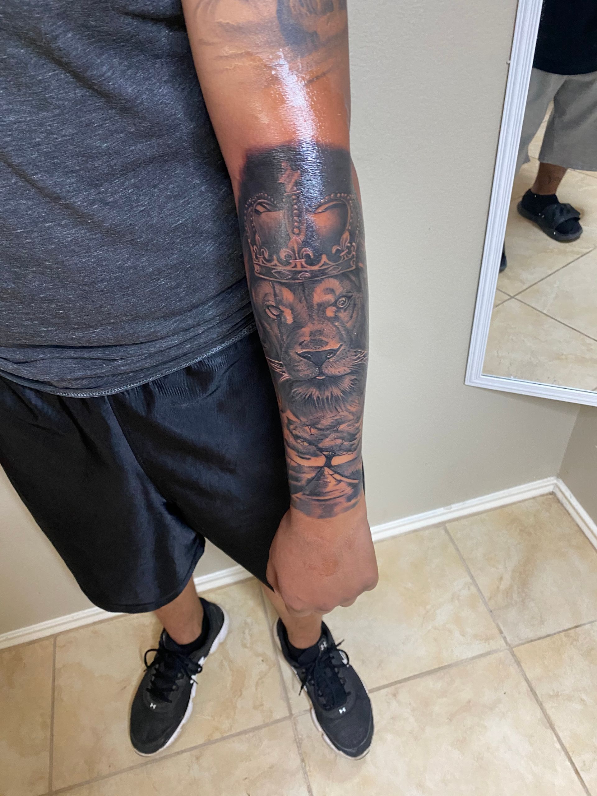 Man with black sleeve tattoo, arm down, wearing black shorts and shoes in a white room.