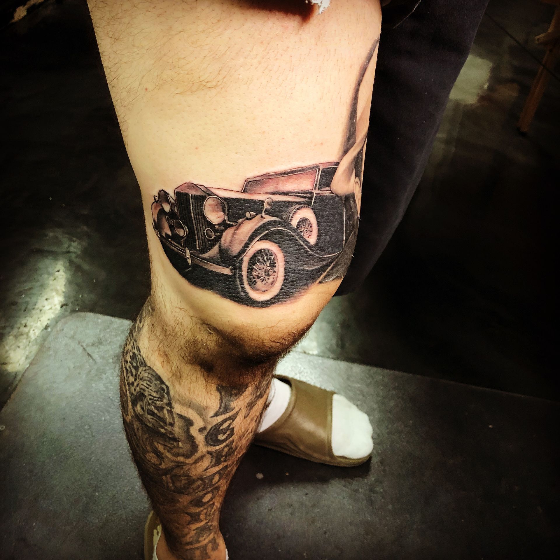 Tattoo of a vintage car on a person's knee. The leg has additional tattoos.
