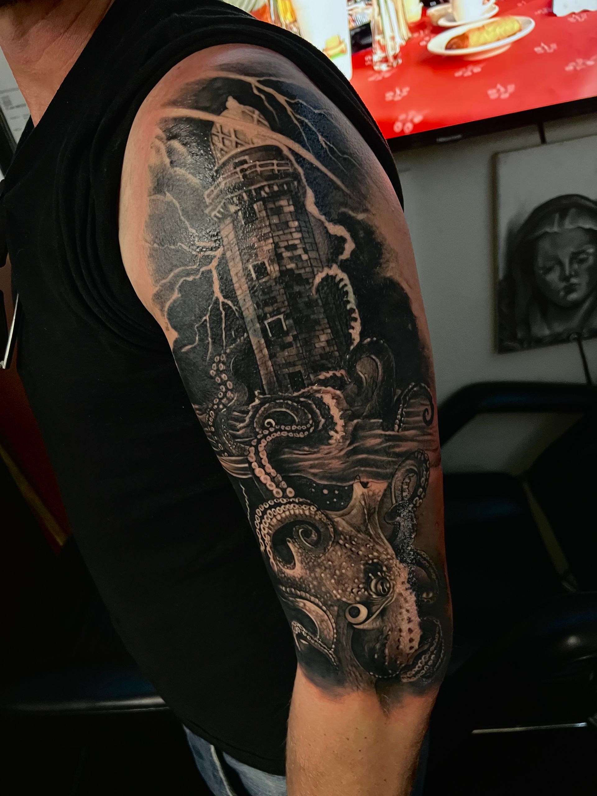Black and grey tattoo of a tower, octopus tentacles, stormy waves on a man's upper arm.