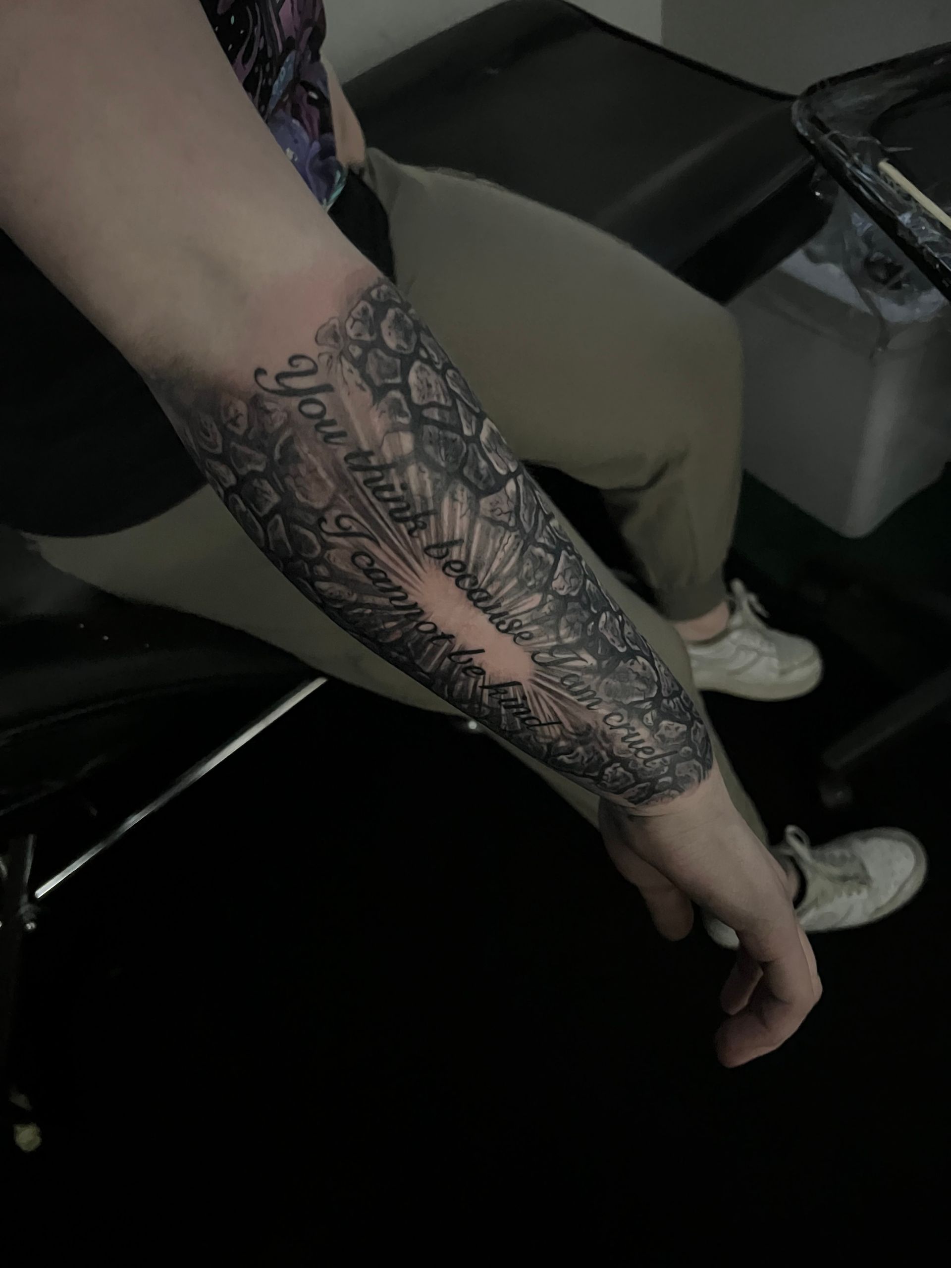 Tattoo of a gray floral design with lettering on a person's forearm. They wear gray pants and white shoes.