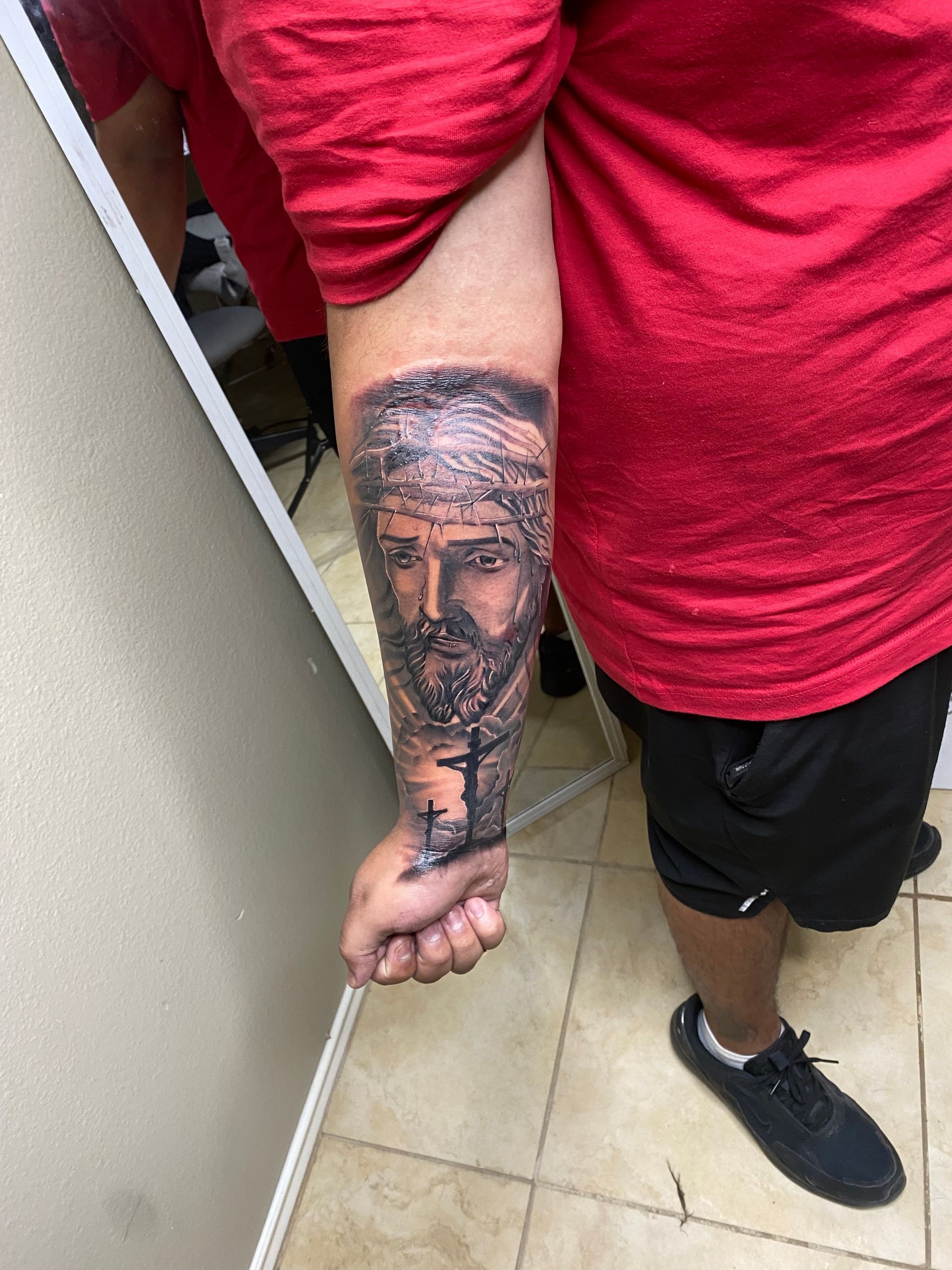 Person with red shirt and black shorts, shows a forearm tattoo of a bearded man with a cross.