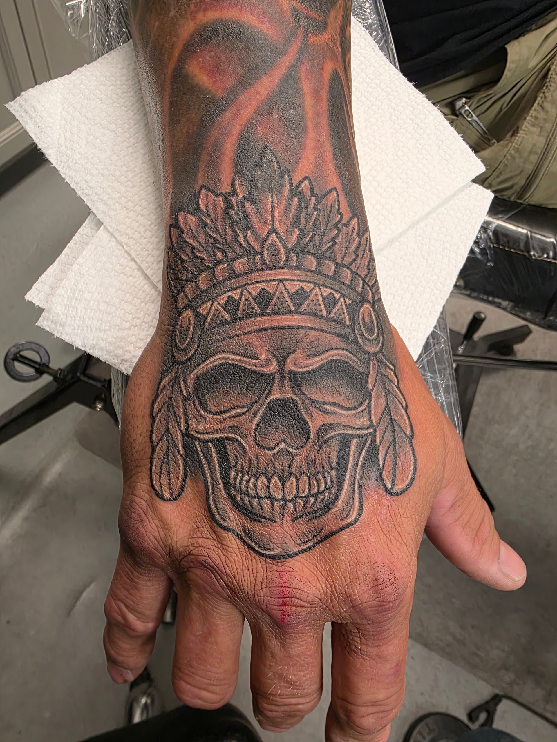 Skull tattoo on hand, wearing a Native American headdress. Black and grey ink.