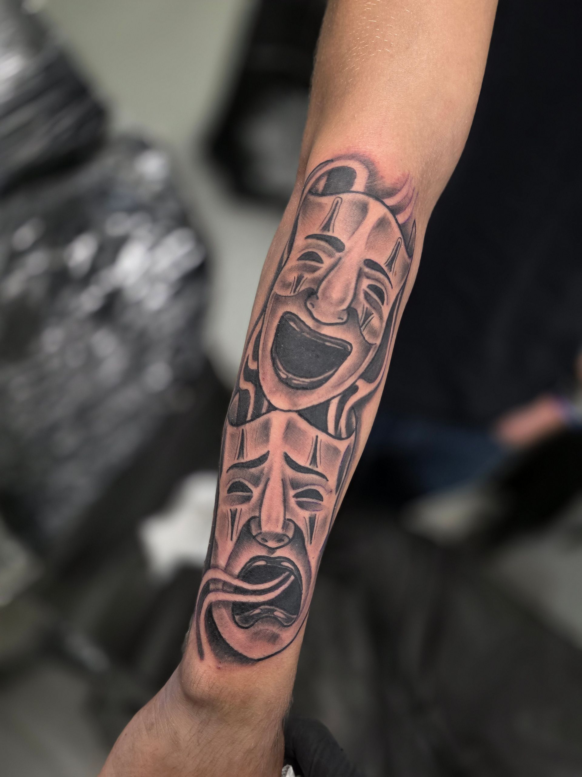Black and gray tattoo of comedy and tragedy masks on a forearm. The masks are stacked vertically.