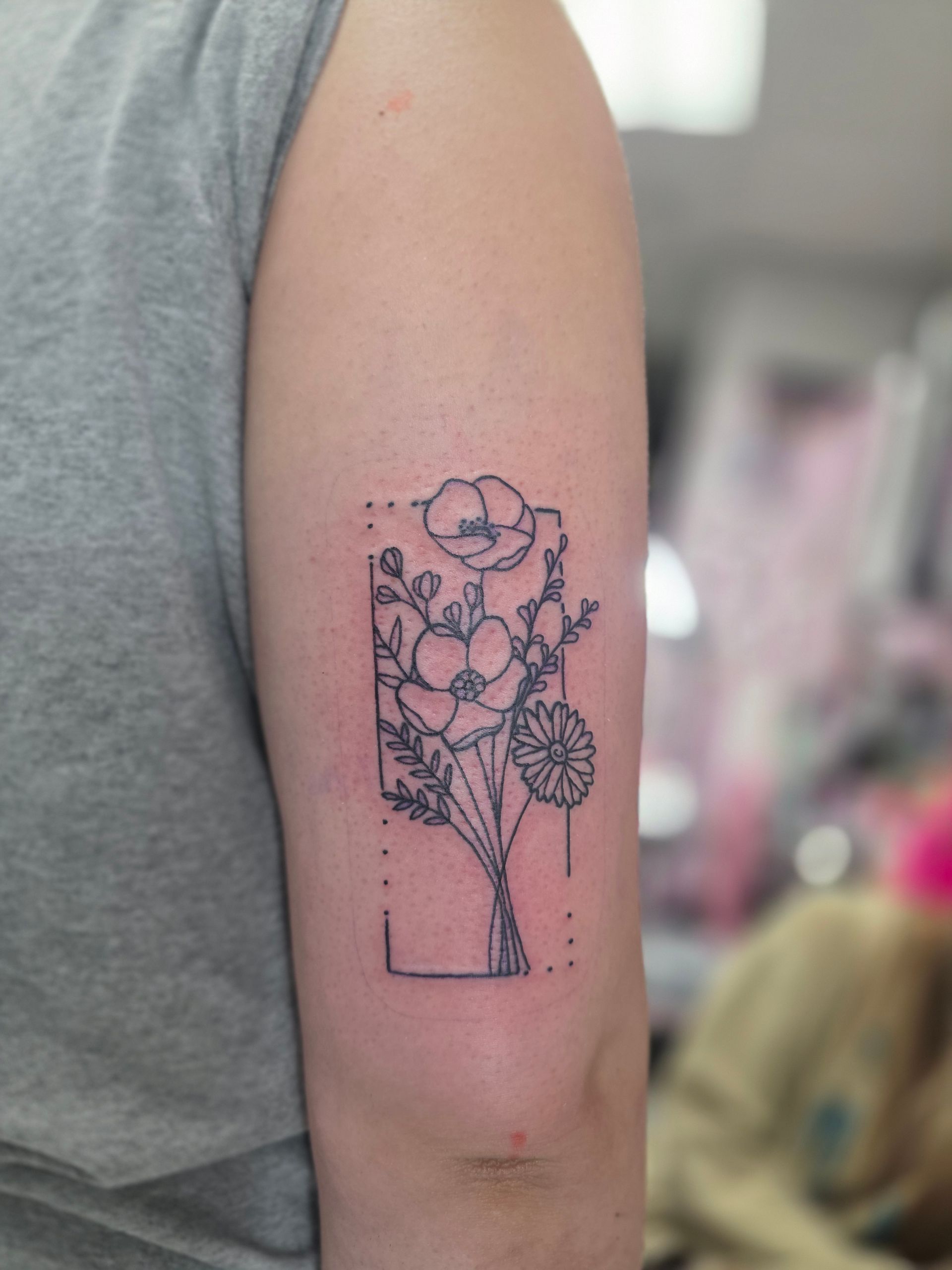 Tattoo of a floral bouquet in a rectangular frame, inked on a person's upper arm.