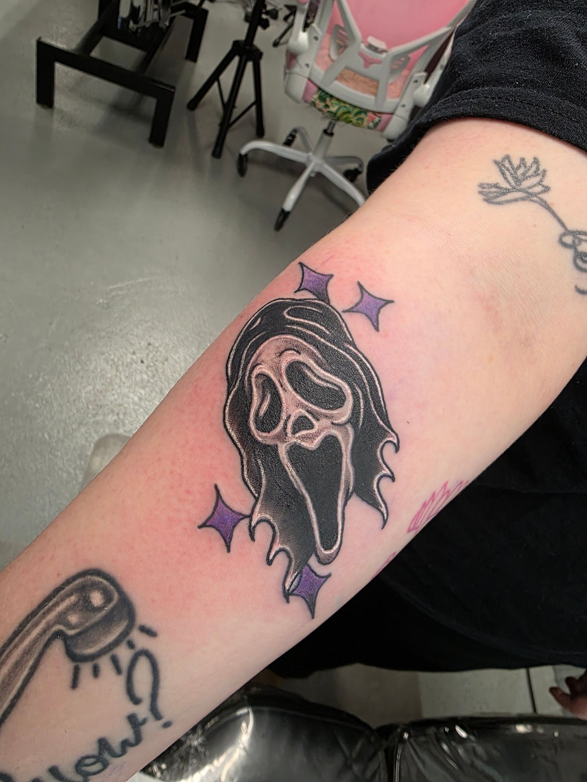 Tattoo of Ghostface mask with purple stars on arm, next to a showerhead tattoo.