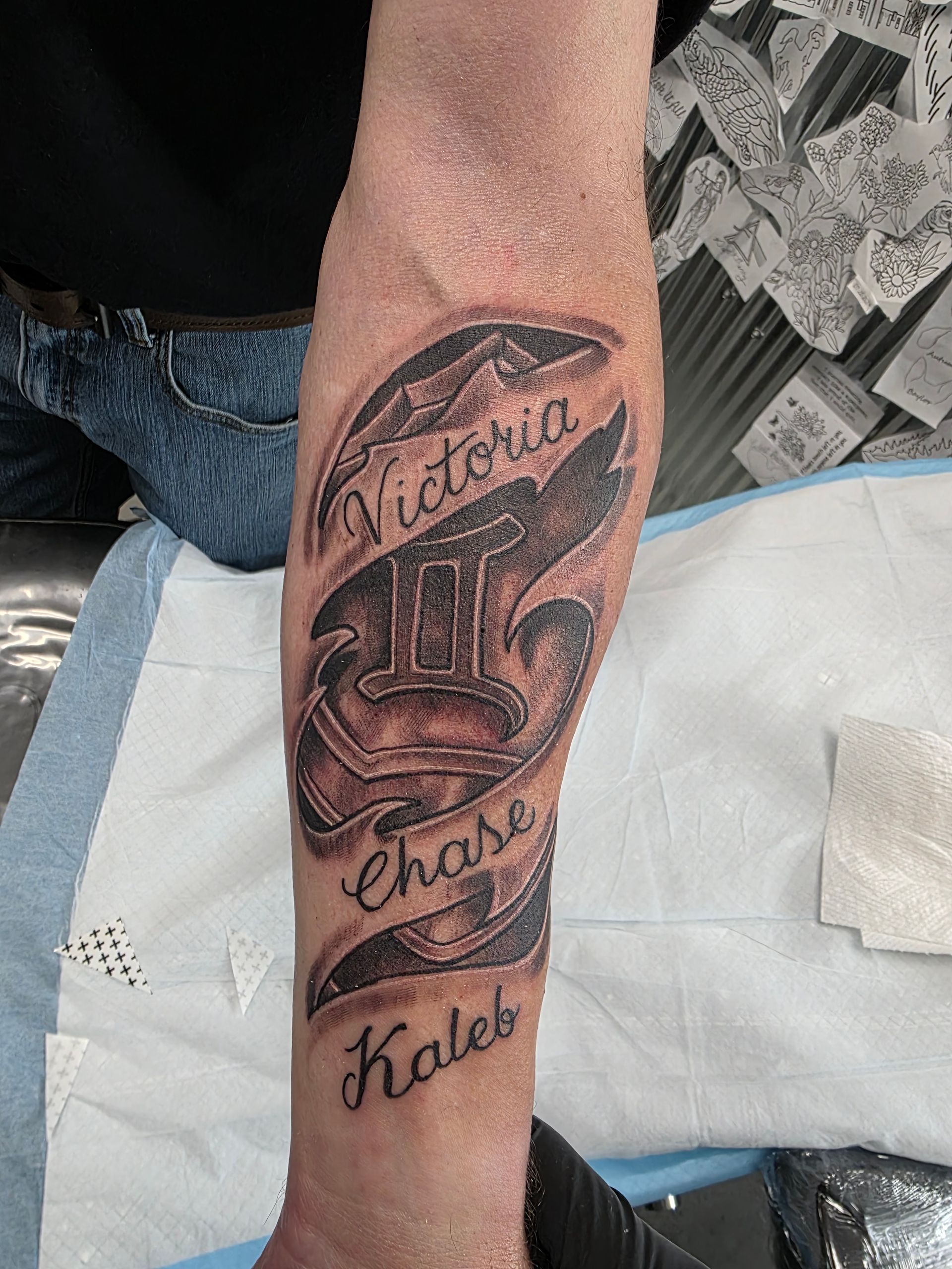 Tattoo of the Gemini symbol with the names Victoria, Chase, and Kaleb on a person's arm.