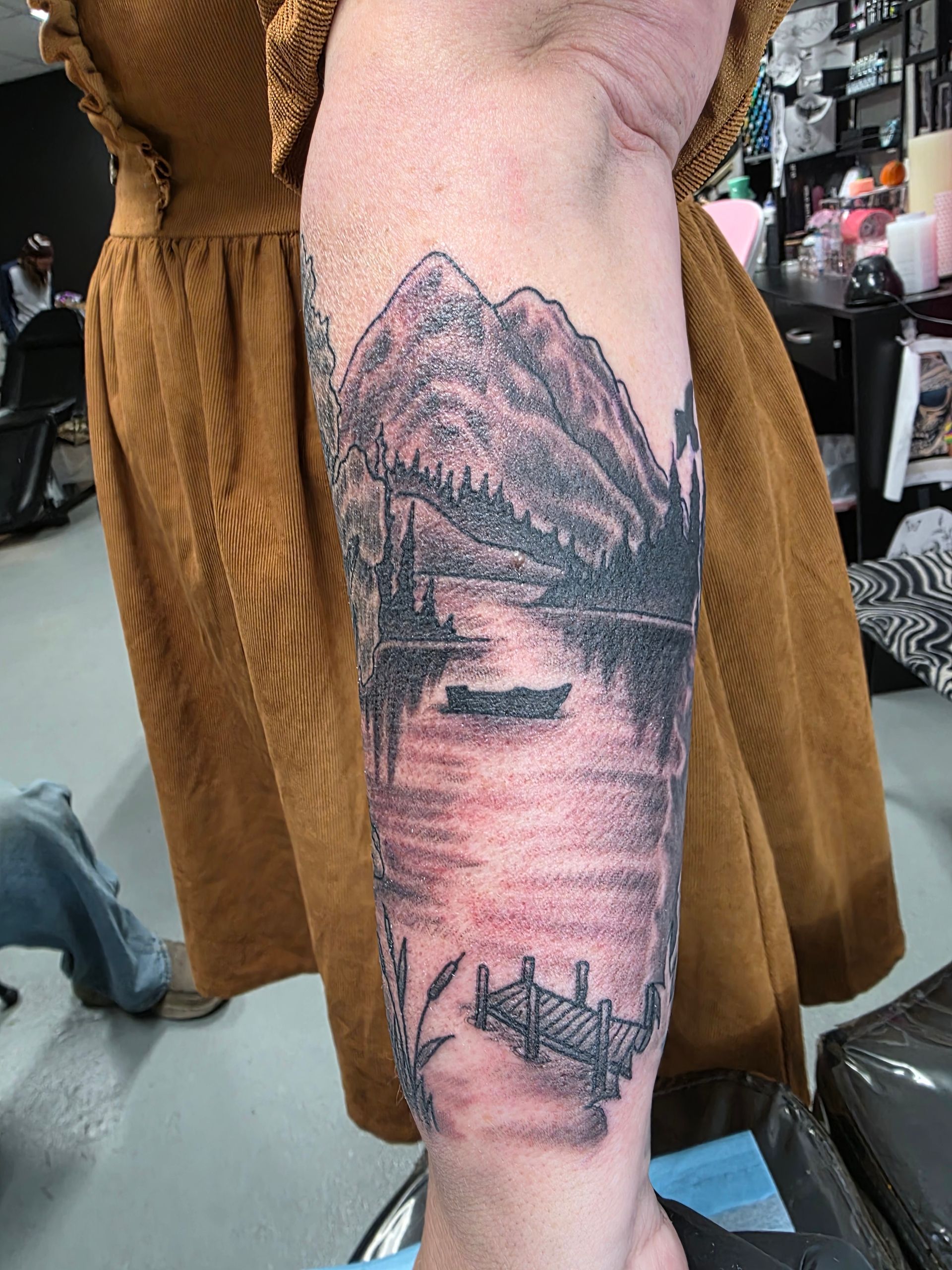 Tattoo of a mountain scene on a person's forearm. Features a lake, boat, pier, and trees in gray ink.