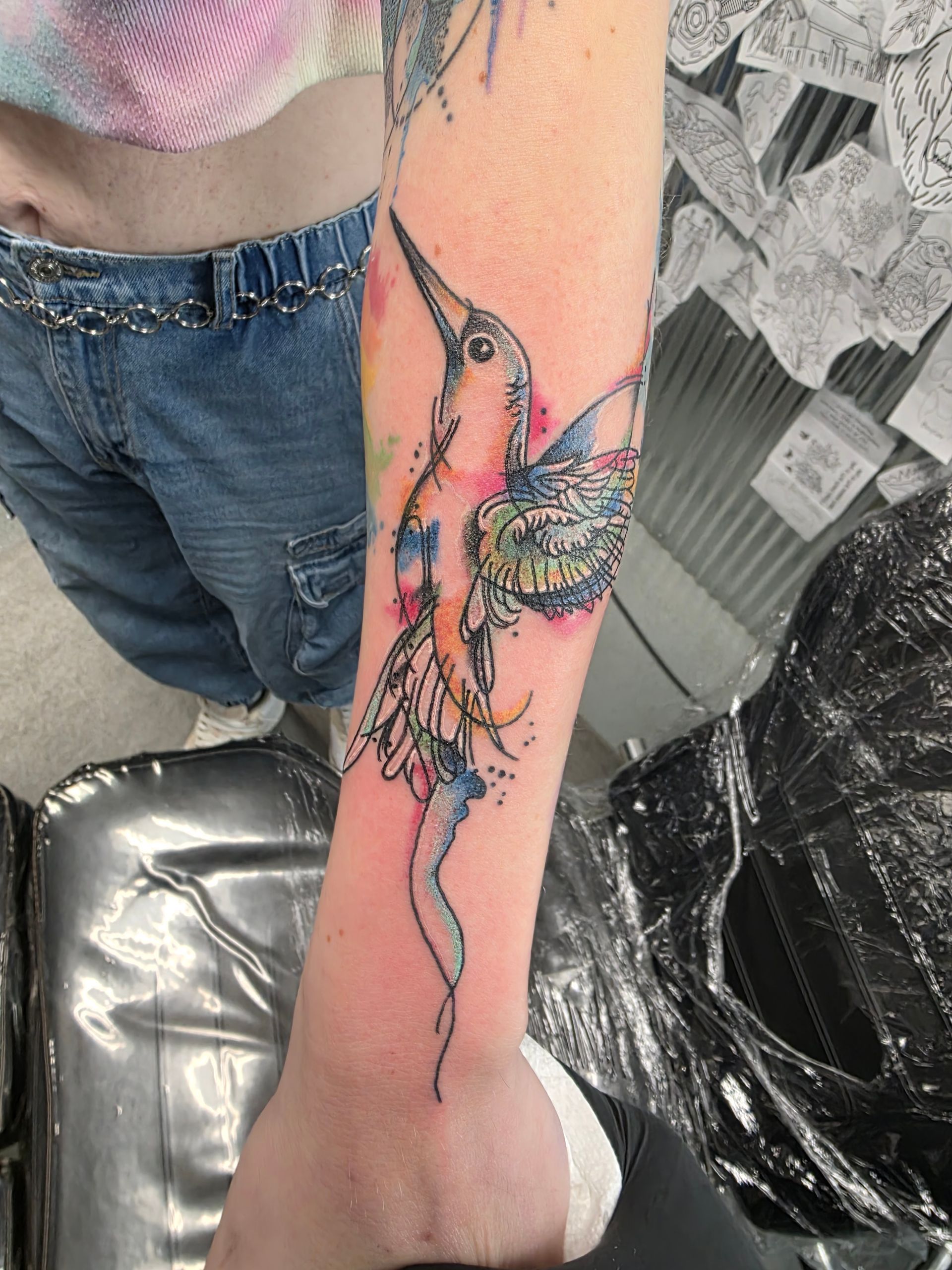 Tattoo of a watercolor-style hummingbird on a person's forearm. Blues, pinks, and yellows are visible.