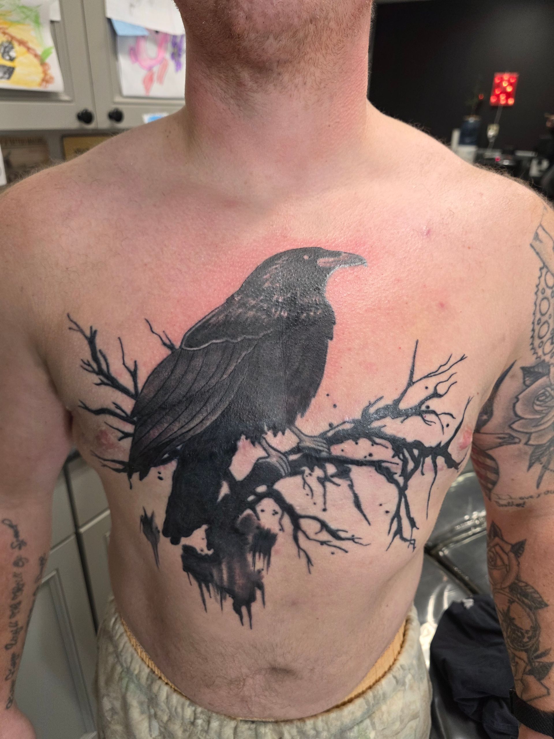 Man with a black raven tattoo on his chest, perched on a tree branch, indoor setting.