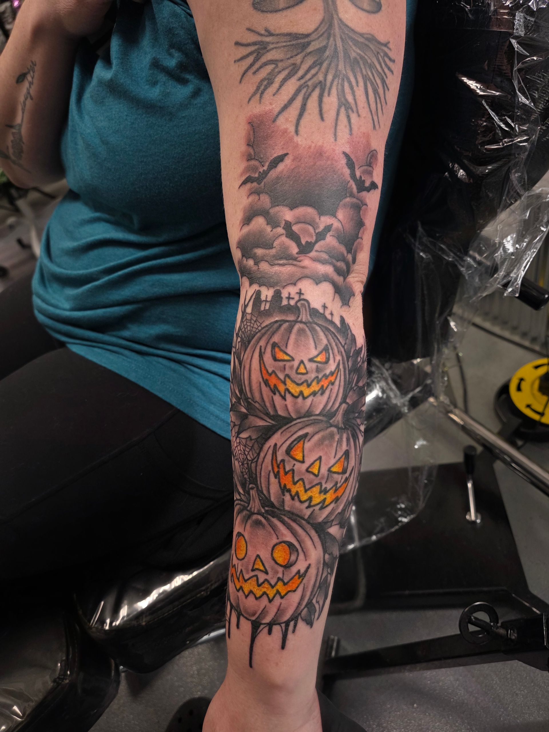 Halloween-themed tattoo of stacked jack-o'-lanterns, bats, and a spooky tree on an arm.