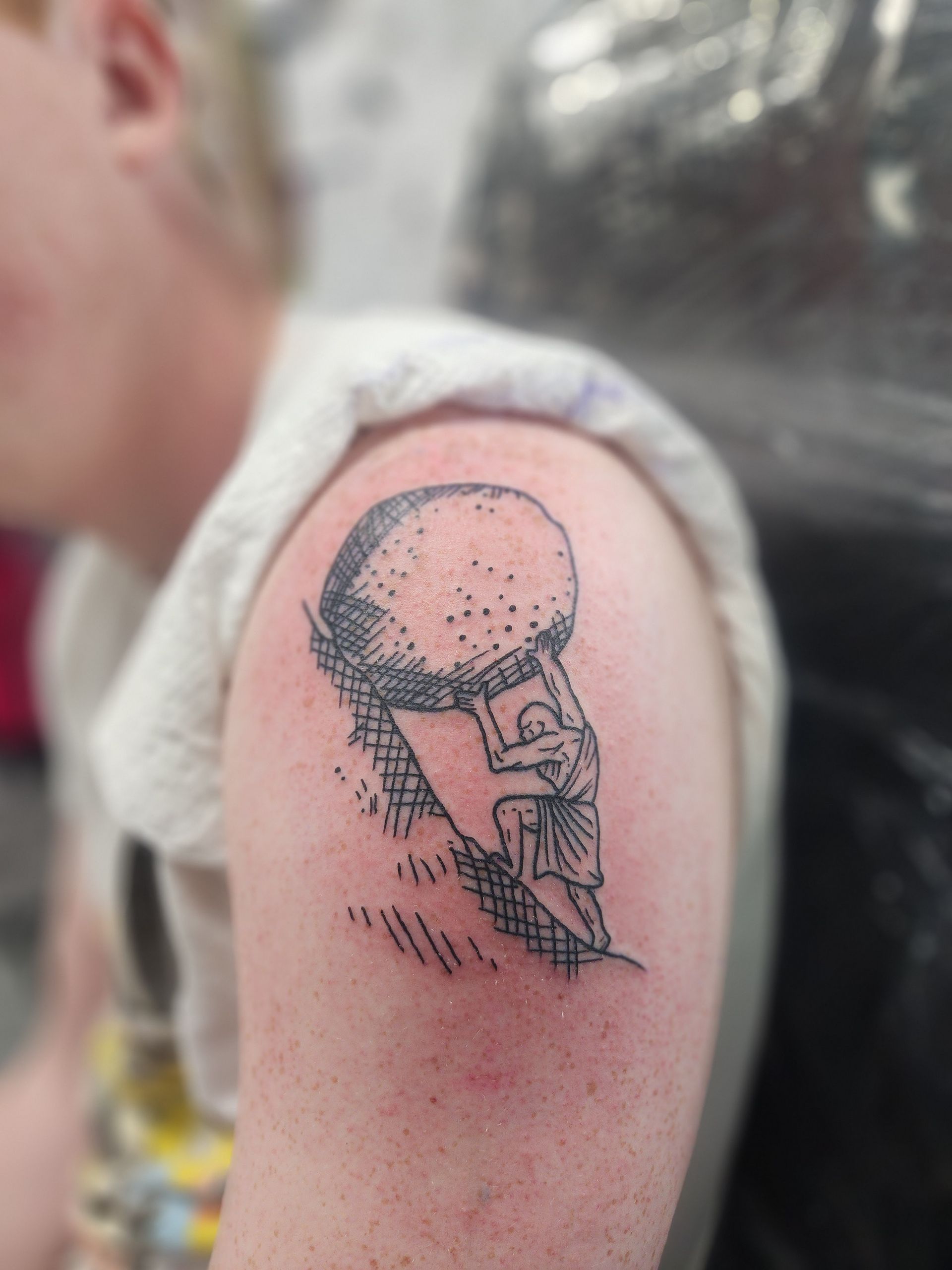 Tattoo of Sisyphus pushing a boulder uphill. Black linework on shoulder.