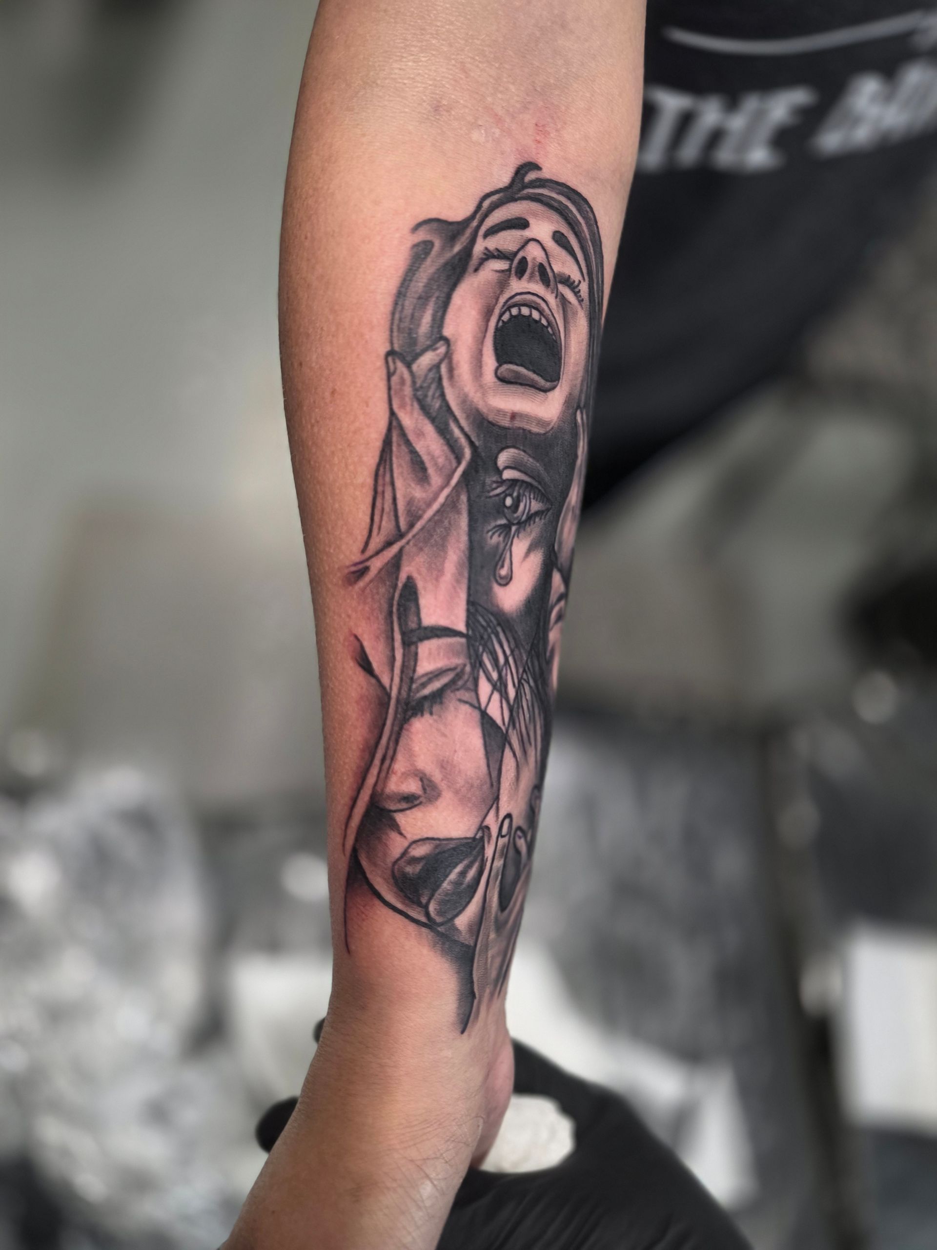 A black and gray tattoo on an arm depicting three faces, one screaming, another with closed eyes.