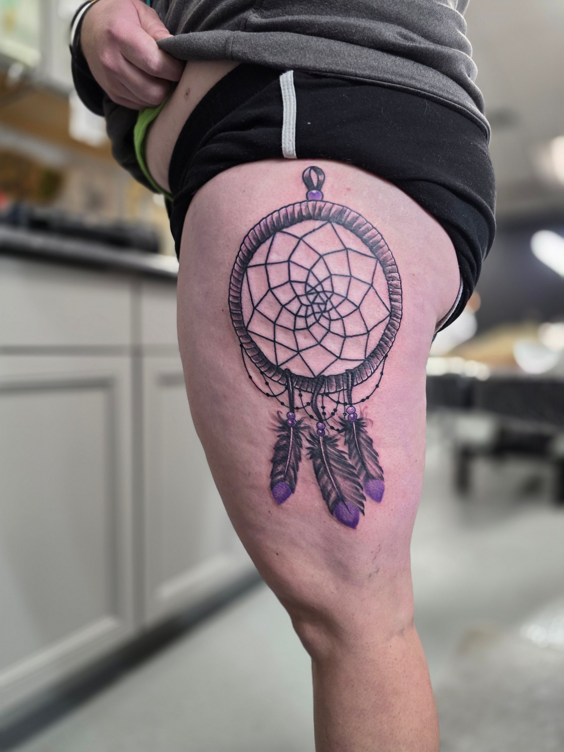 Dreamcatcher tattoo on a person's thigh; black outline, purple feathers, and a blurred background.
