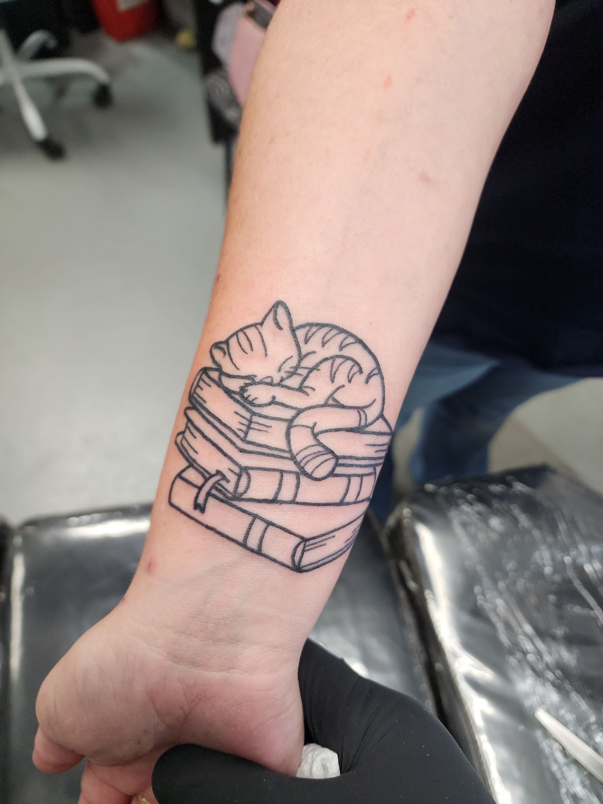 Black-lined tattoo of a sleeping cat on a stack of books on a person's wrist.
