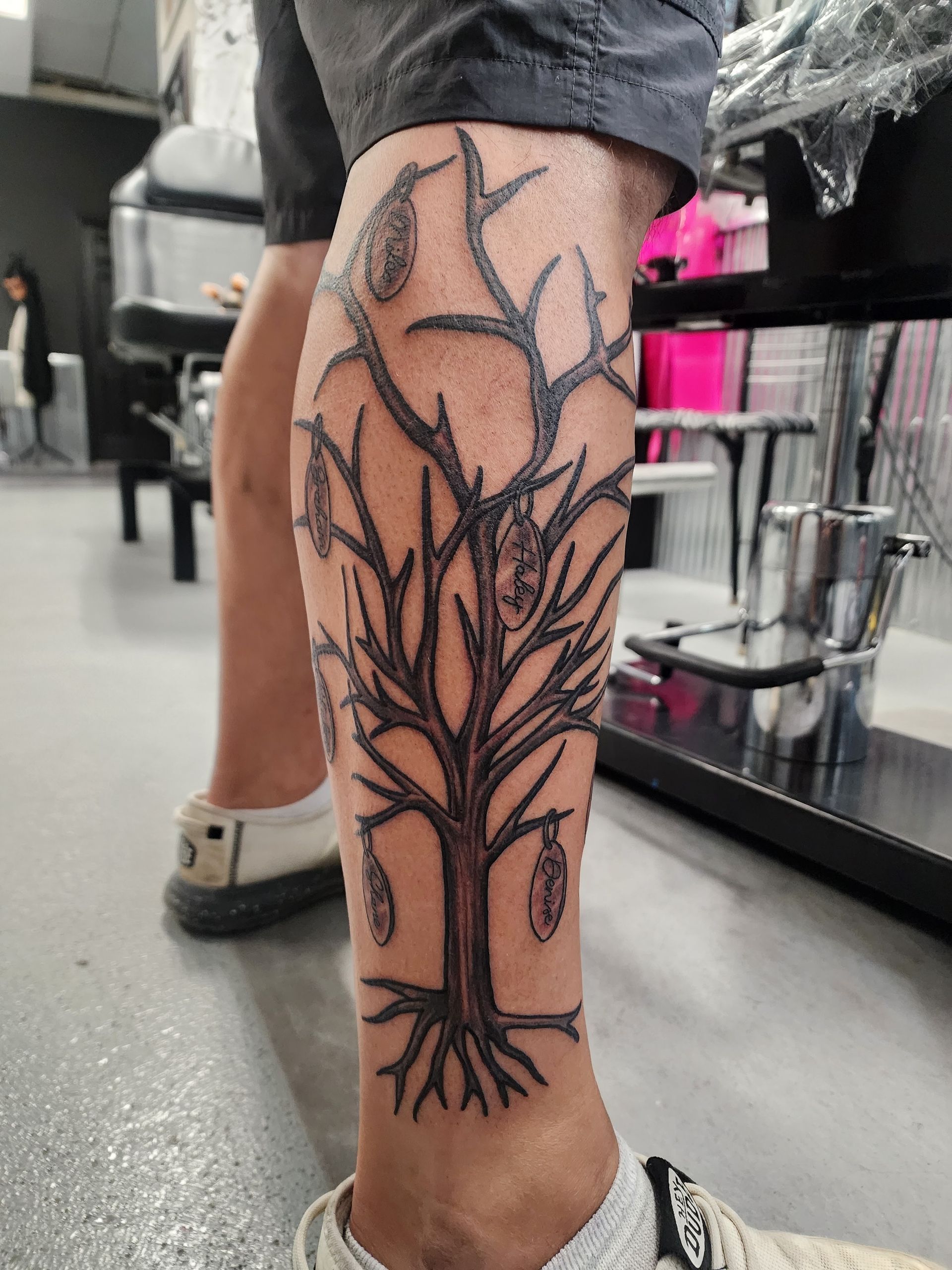 Tattoo of a bare tree on a leg, with leaves/fruit hanging from the branches, in a tattoo parlor.
