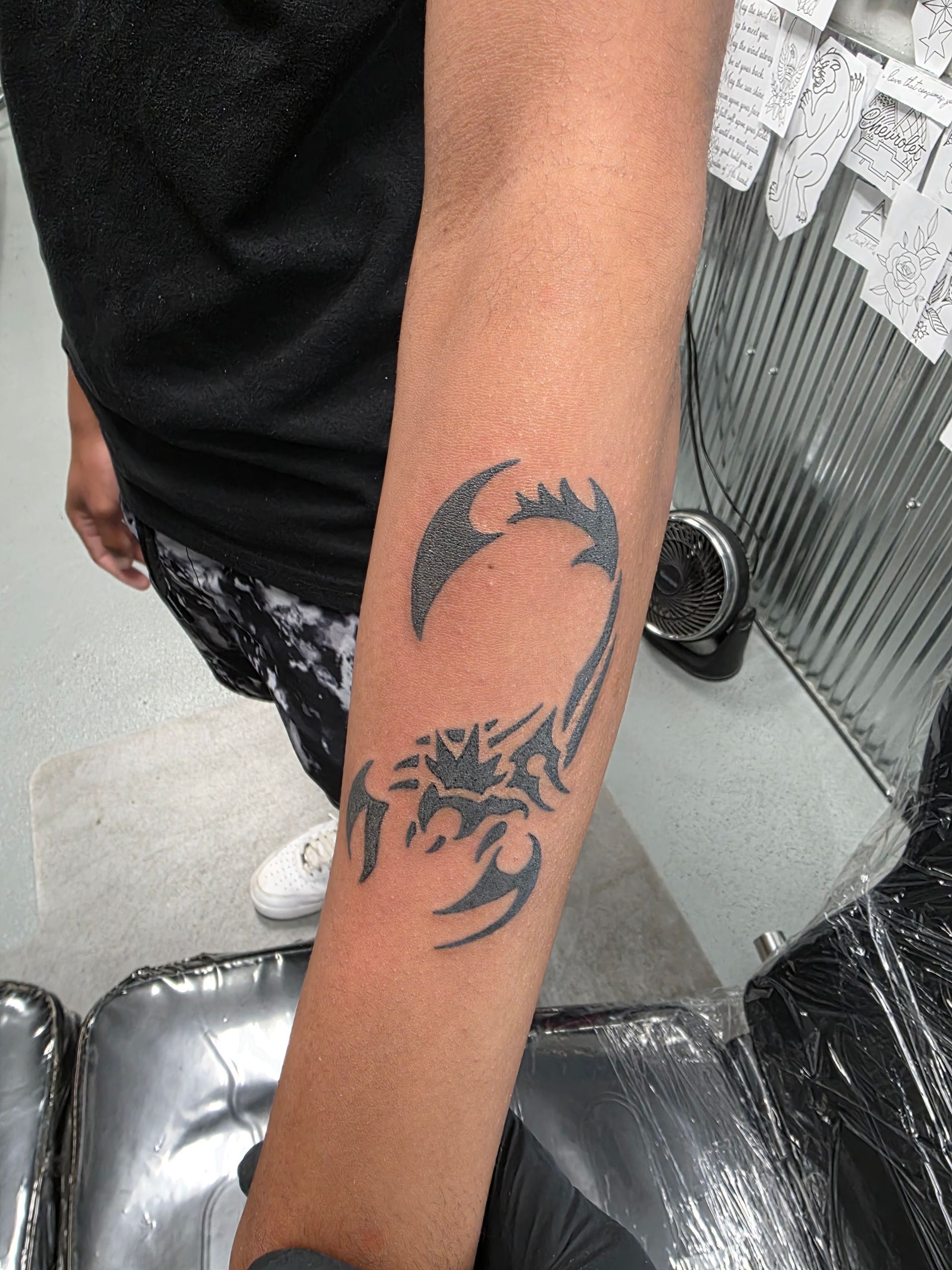 Black scorpion tattoo on forearm. Person wearing black shirt and white shoes.