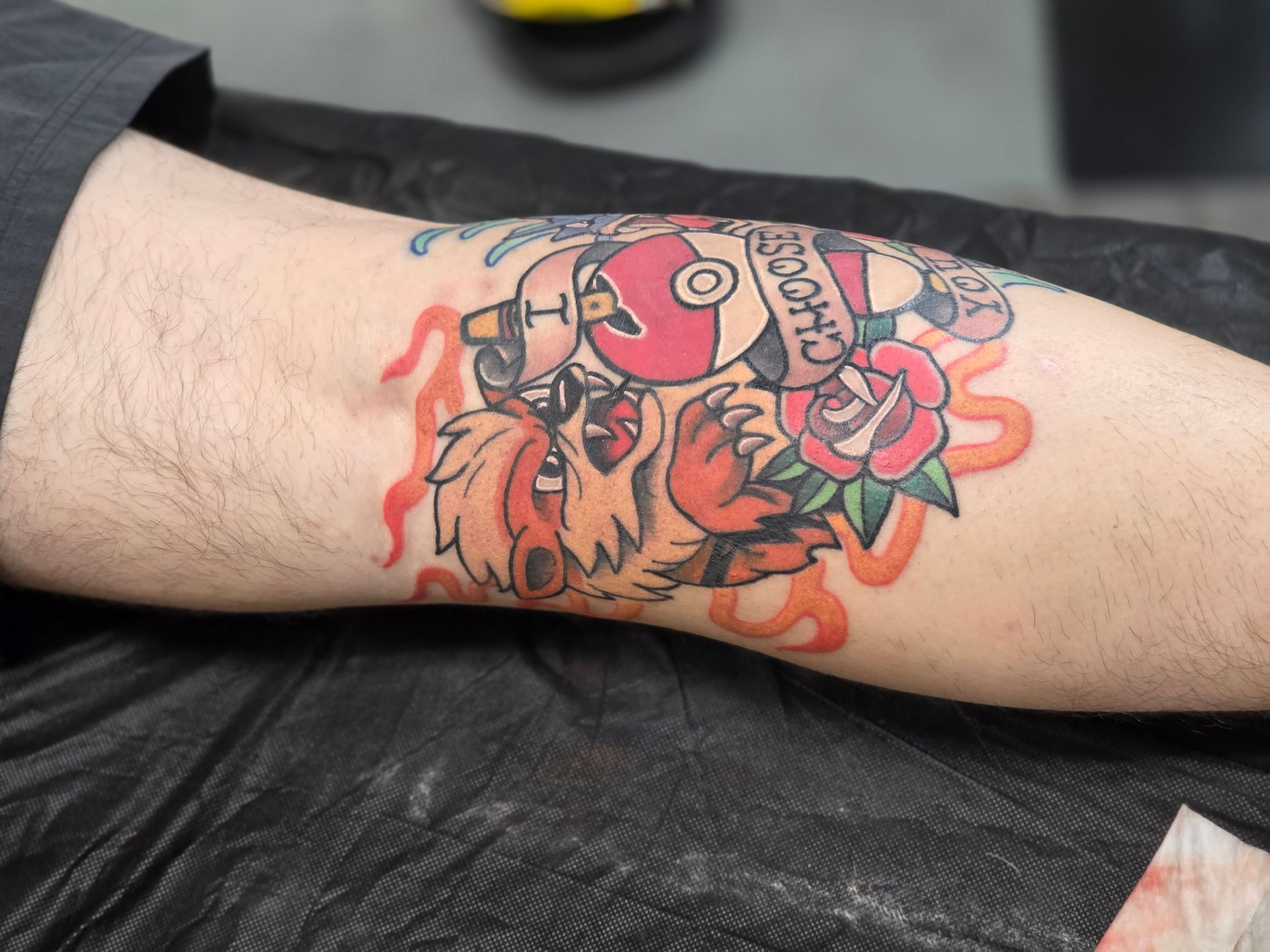 Tattoo of a cartoonish Arcanine, skateboard, and rose on a forearm, surrounded by orange flames and blue accents.