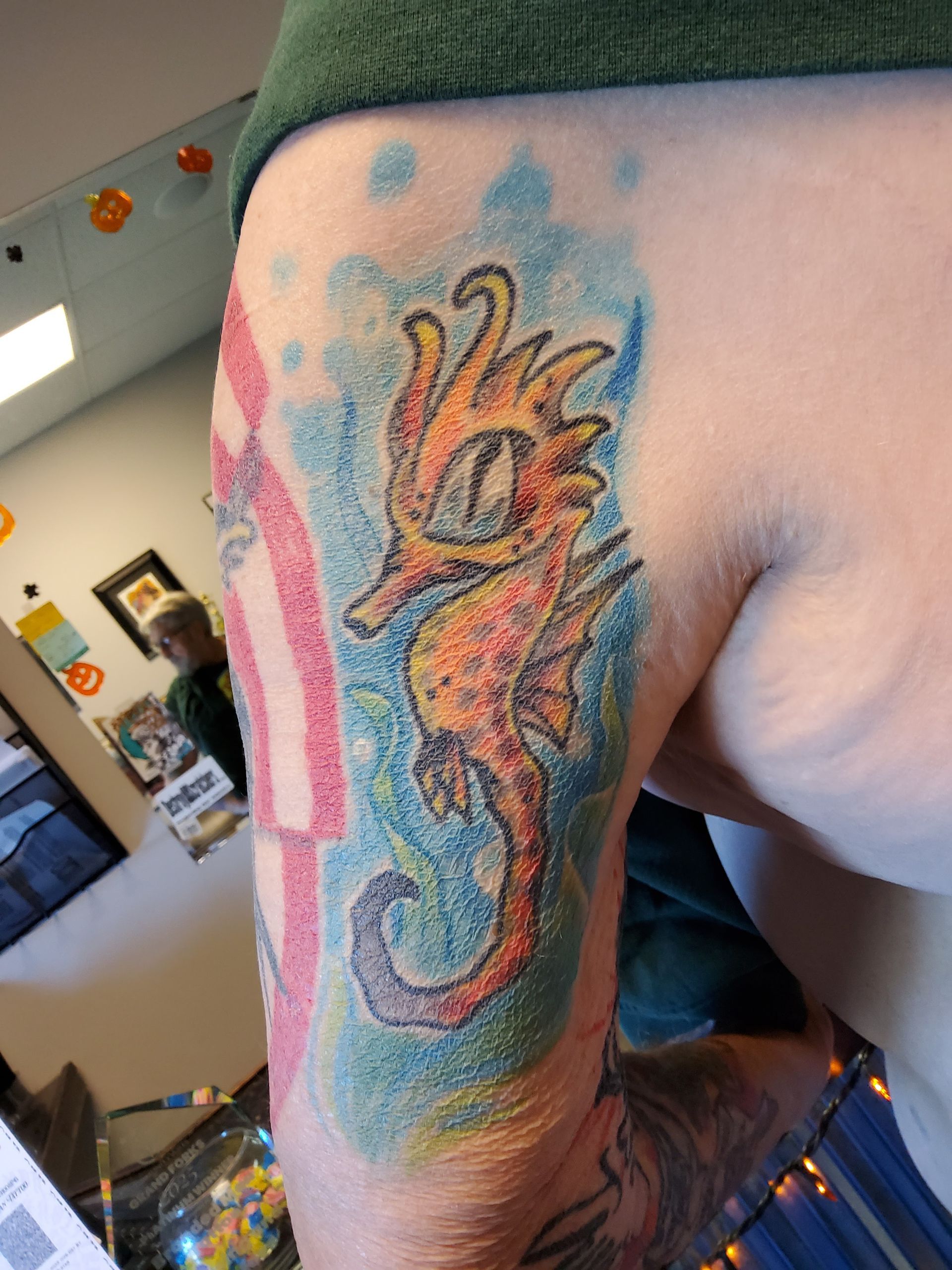 Tattoo of an orange and yellow seahorse with blue, pink, and green watercolor background on a person’s arm.